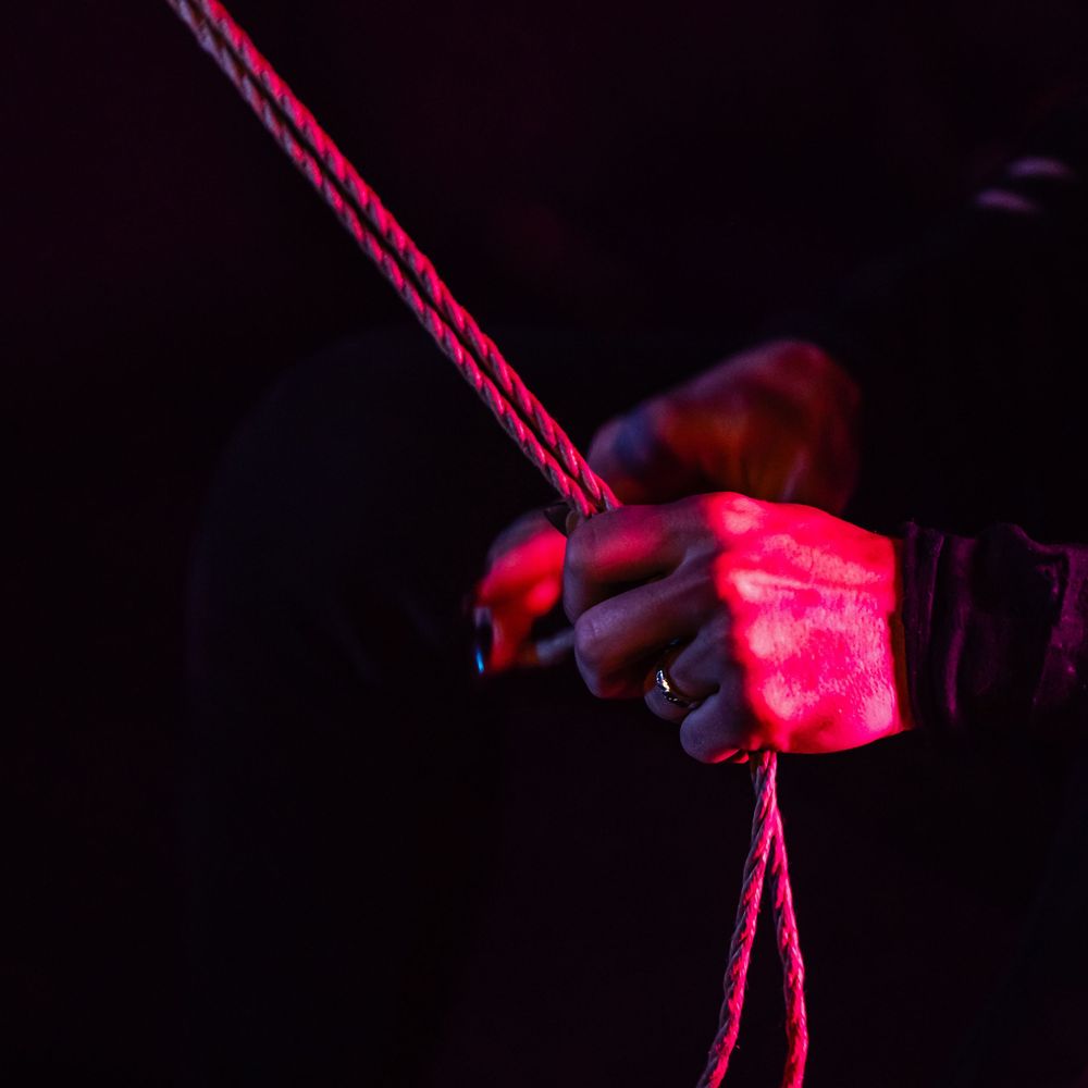 Aerial dancer illuminated in red light performs acrobatic moves on a suspended rope against dark backdrop.