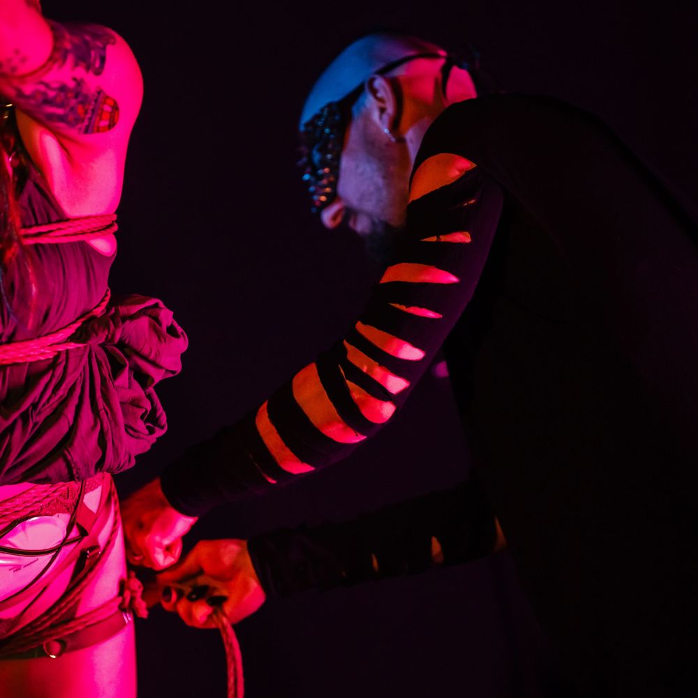 Performer executes dramatic aerial rope choreography bathed in vibrant red and blue stage lighting.