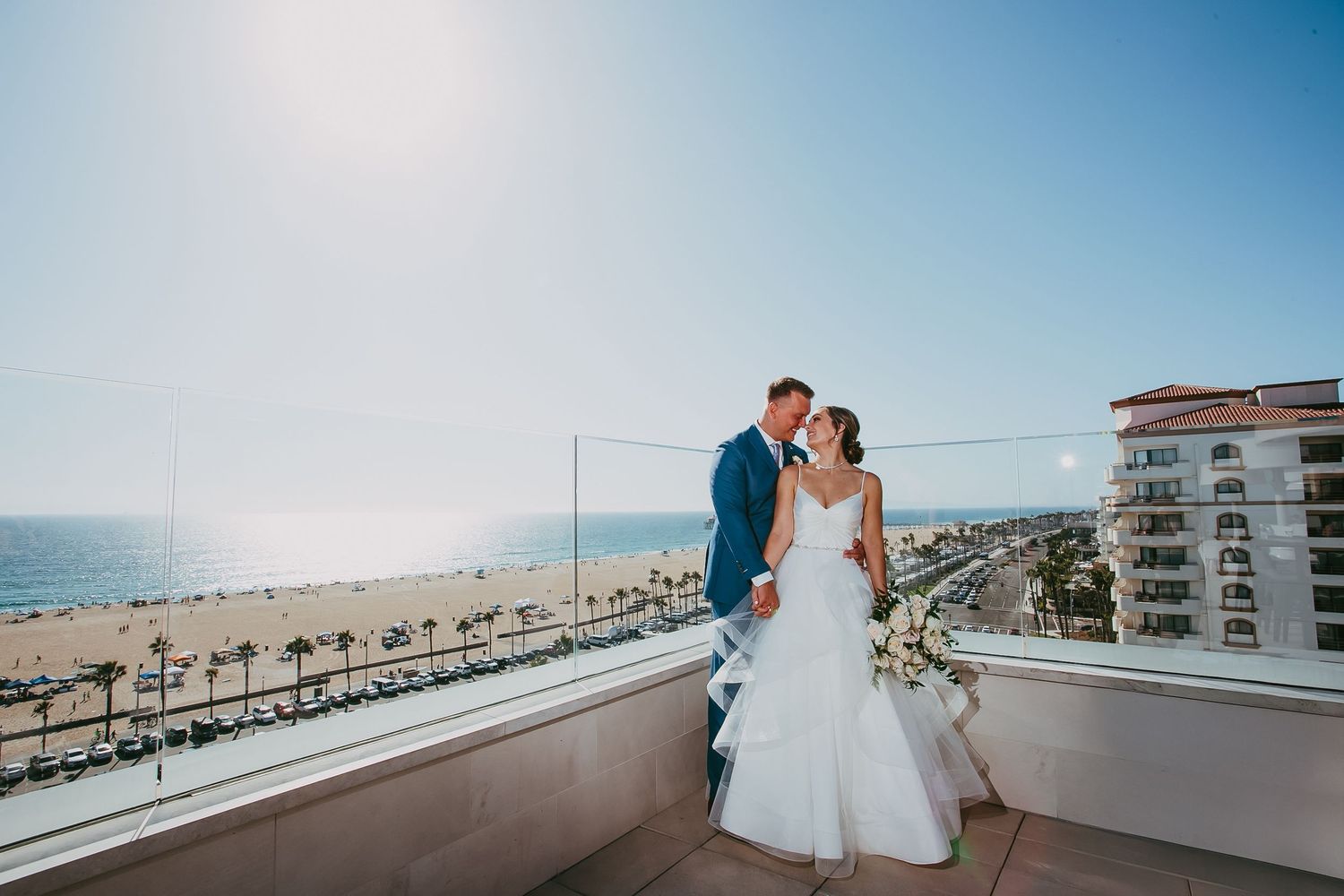 A romantic wedding couple shares a tender moment on a rooftop overlooking a sunny beach coastline in Huntington Beach, California.