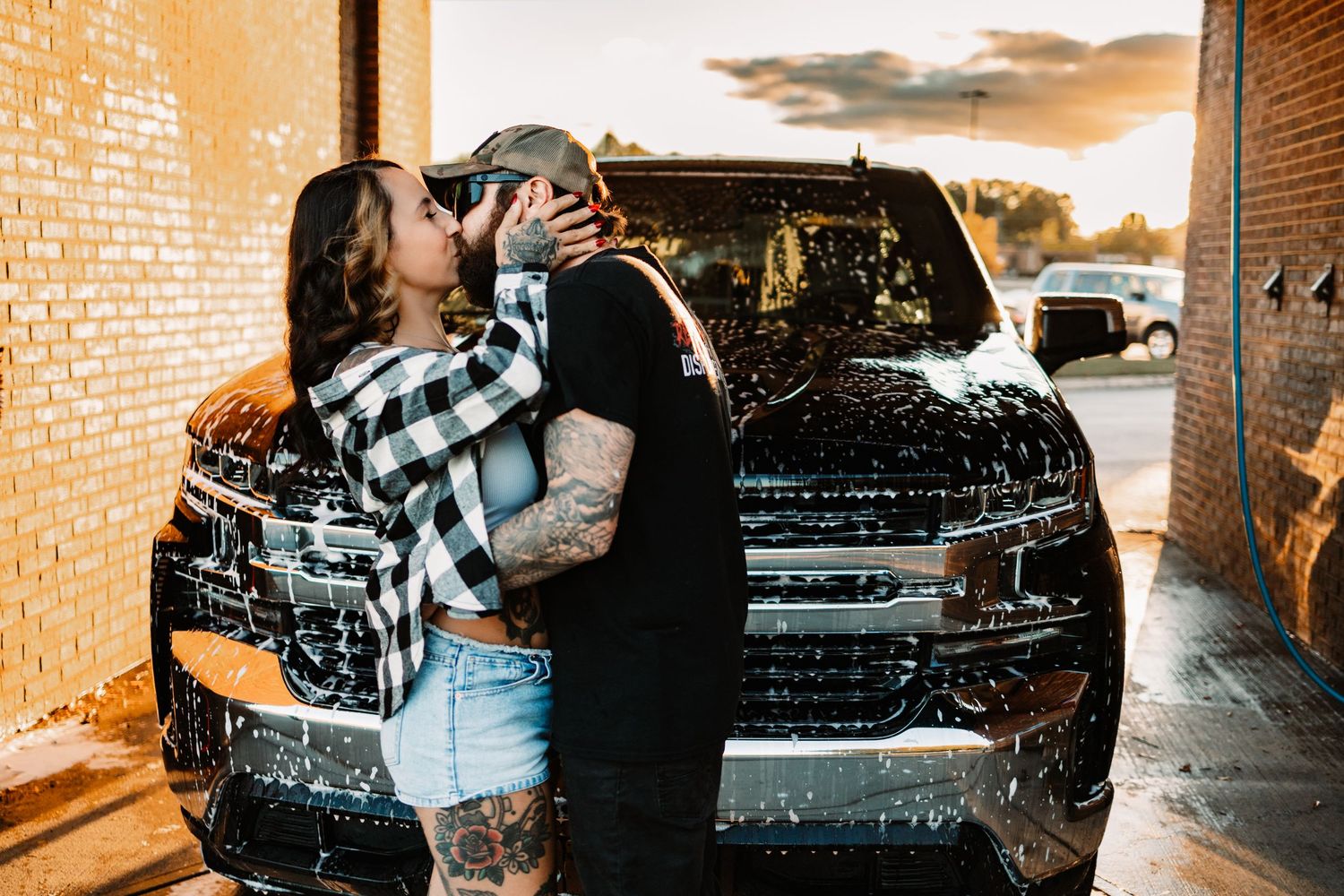 Tattooed couple sharing a romantic kiss in front of a soapy black truck at a self-serve car wash at sunset.