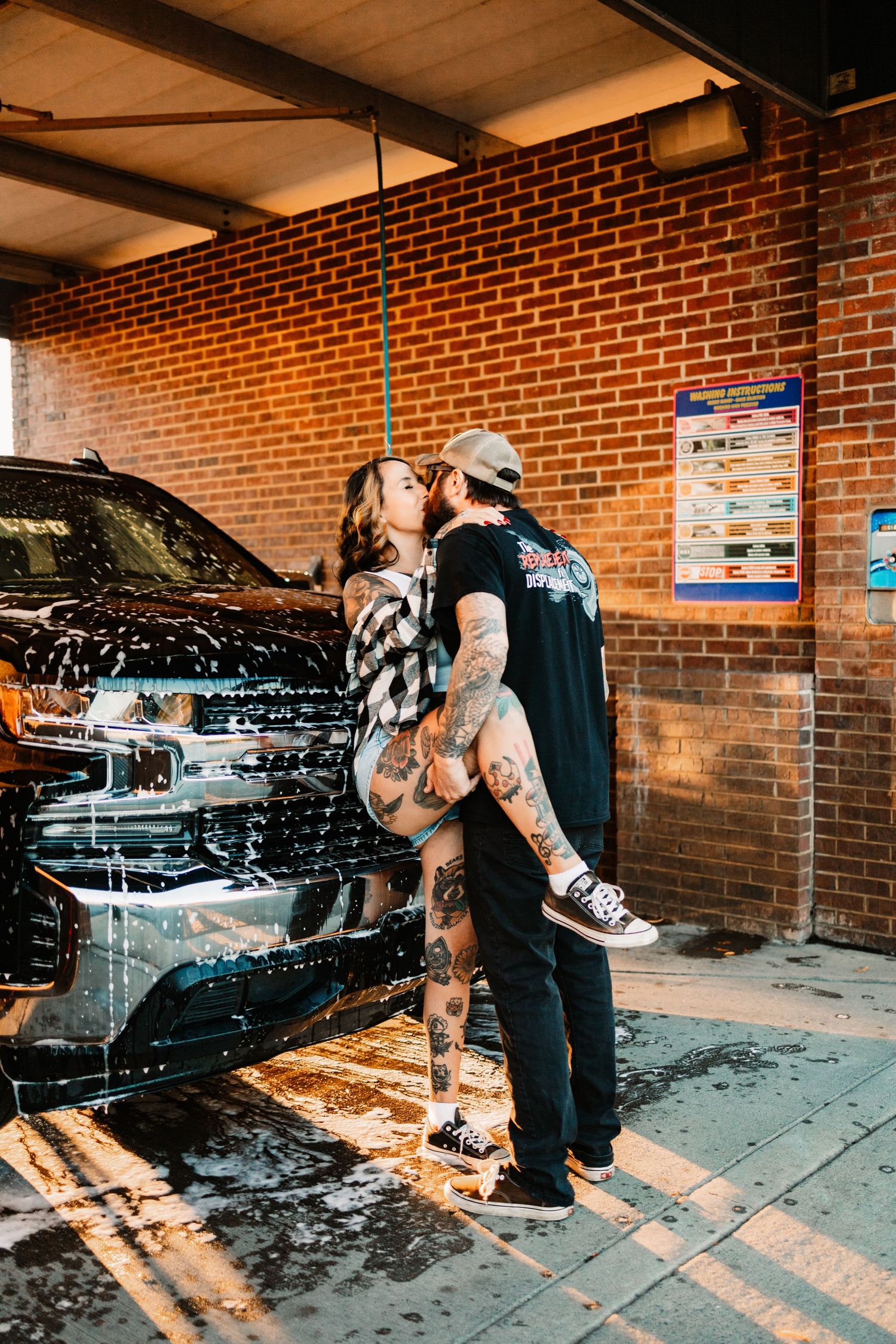 Tattooed man lifting woman for a kiss beside a foam-covered black truck at a brick car wash at golden hour.