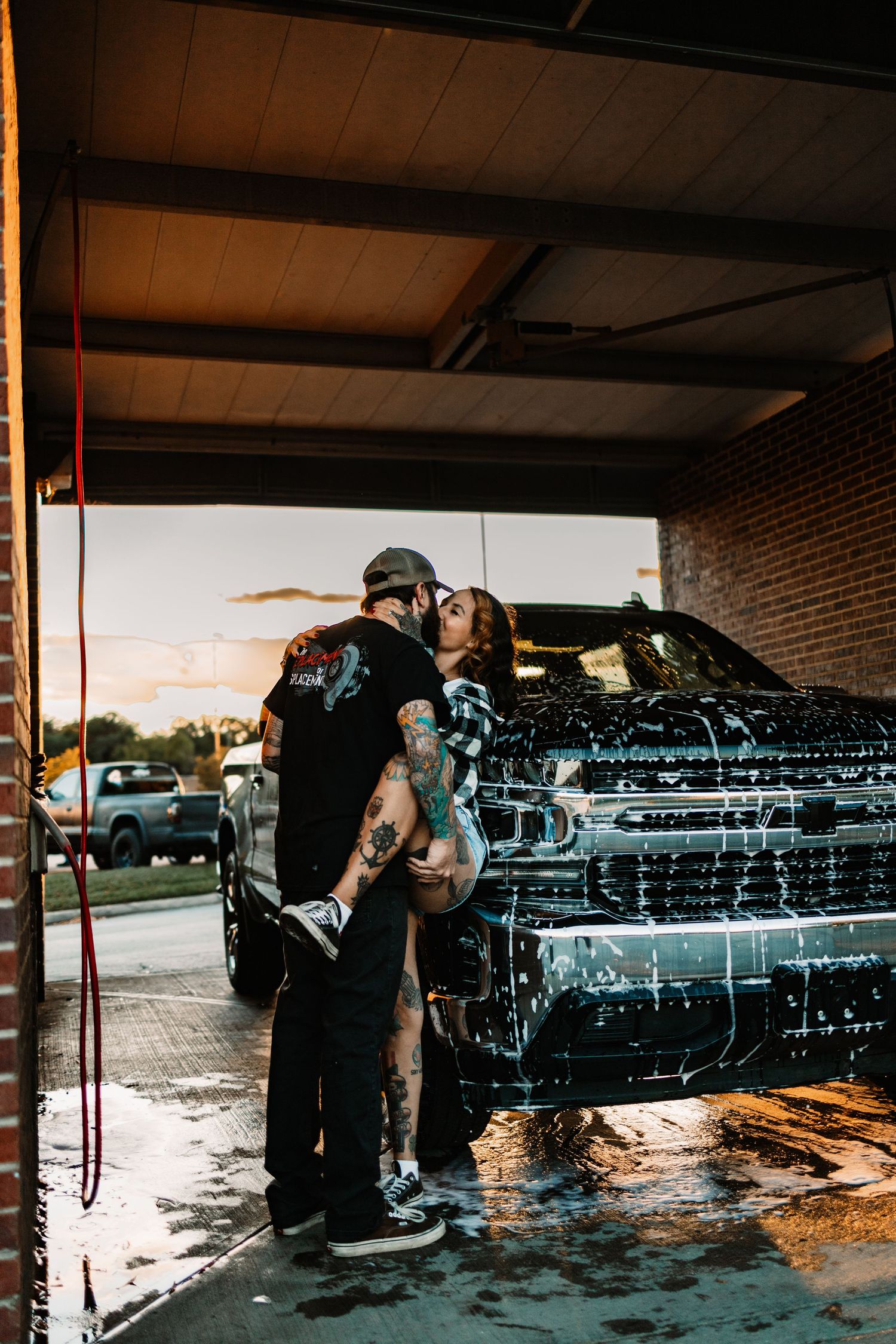 Tattooed couple embracing and kissing beside a soapy black pickup truck at an outdoor car wash at dusk.