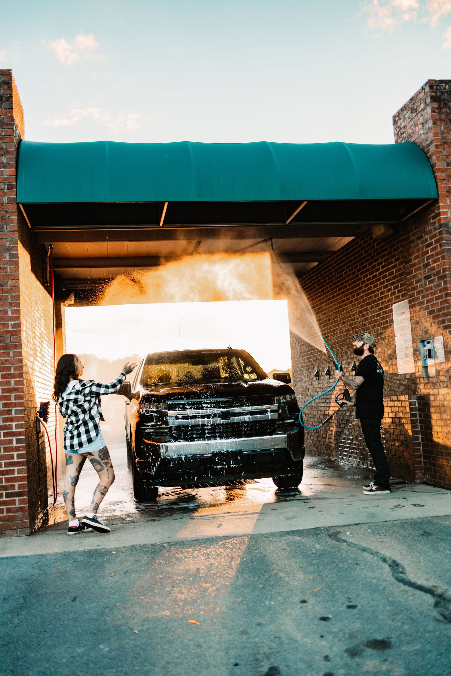 Tattooed couple playfully washing a black truck with pressure hoses at a brick self-serve car wash at sunset.