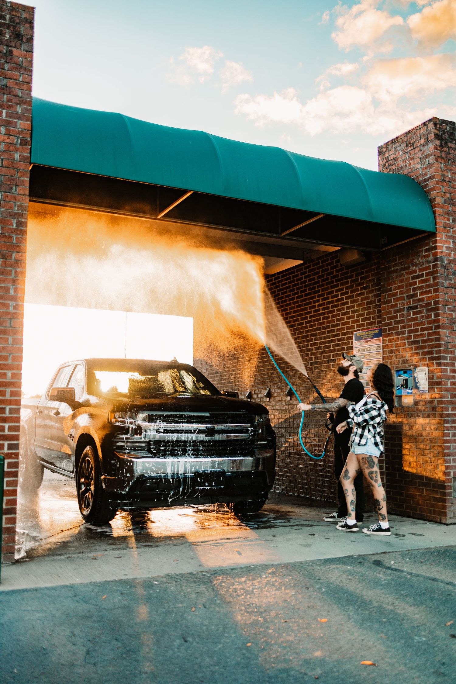 Tattooed woman pressure washing a black pickup truck at a self-serve car wash bay during a golden sunset.