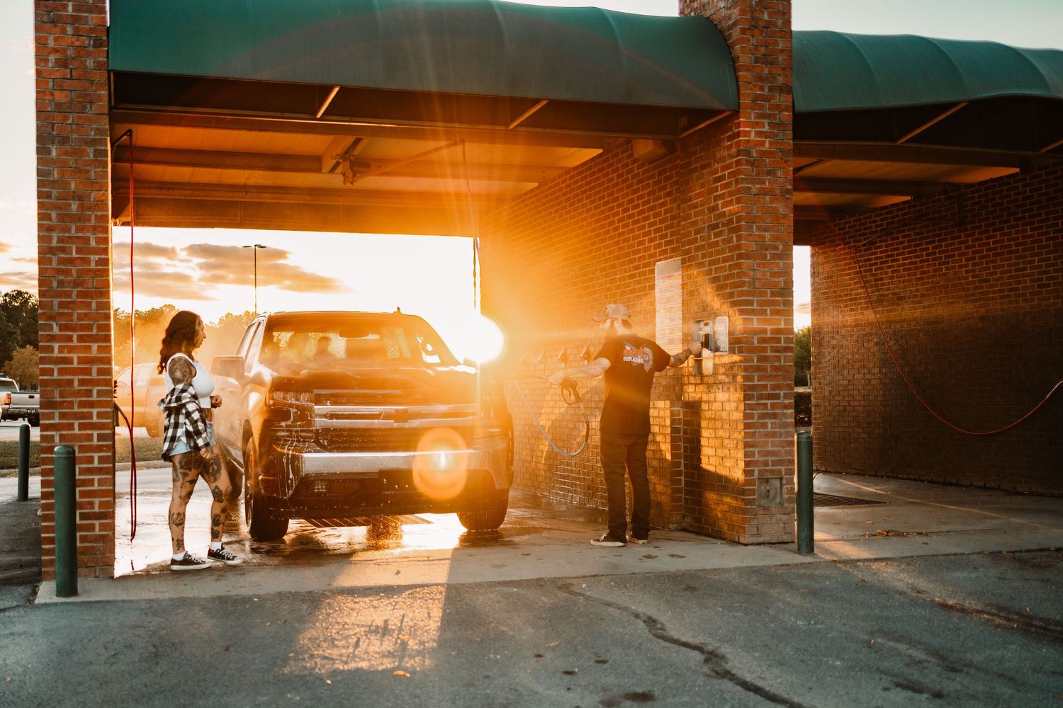 Couple standing beside a black pickup truck at a brick car wash bay bathed in warm golden sunset light.