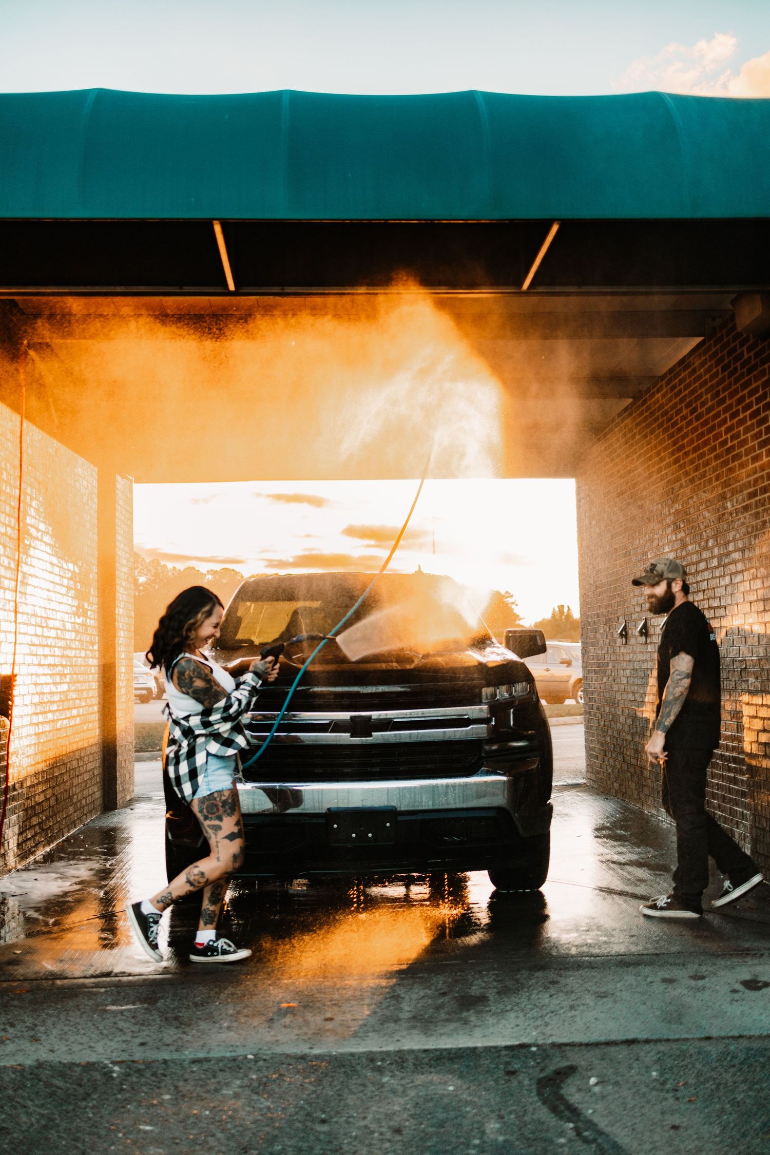 Tattooed woman power washing a black truck while man watches at a self-serve car wash during vivid sunset.