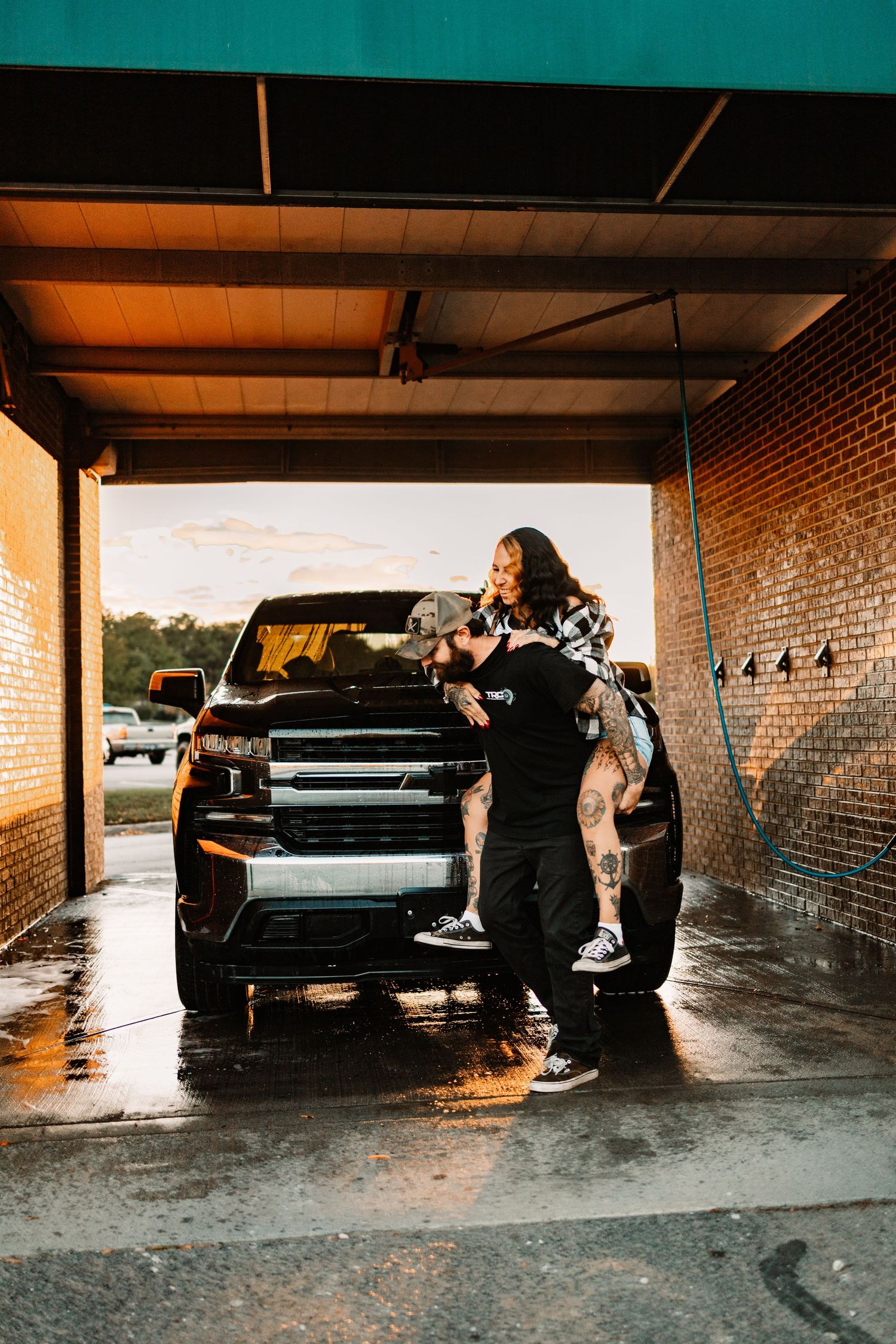 Couple embracing in front of a black SUV at a self-serve car wash bay during golden hour sunset.