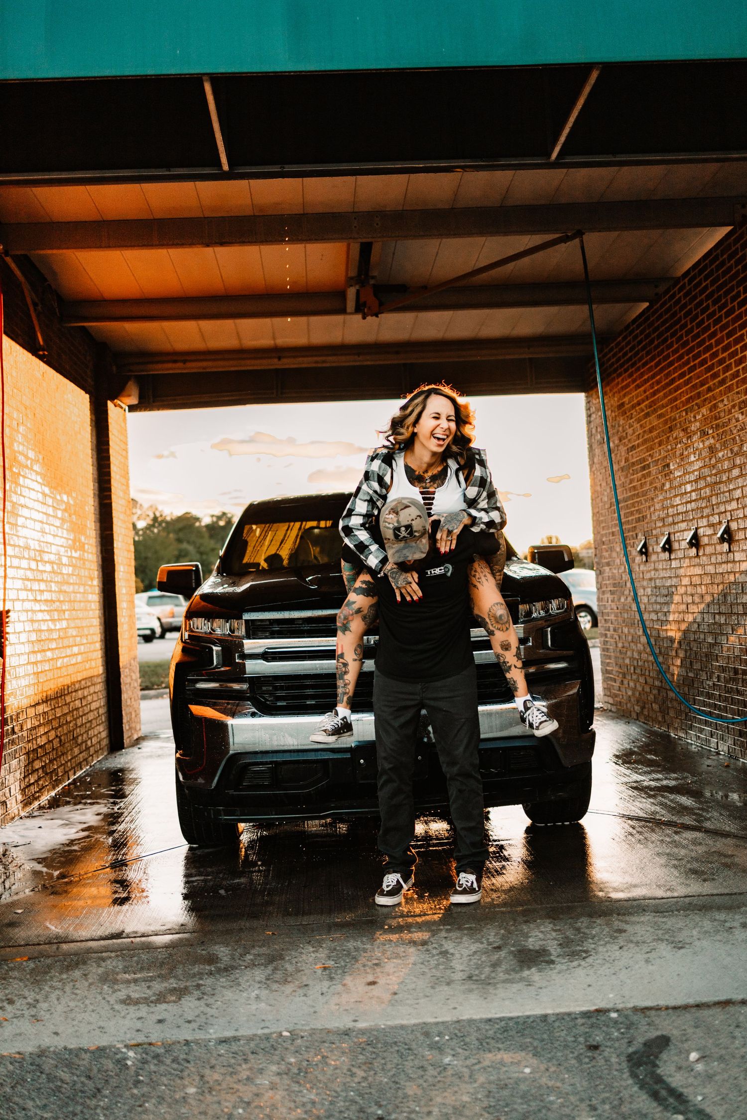 Man carrying laughing woman on his shoulders in front of black truck at car wash during sunset.