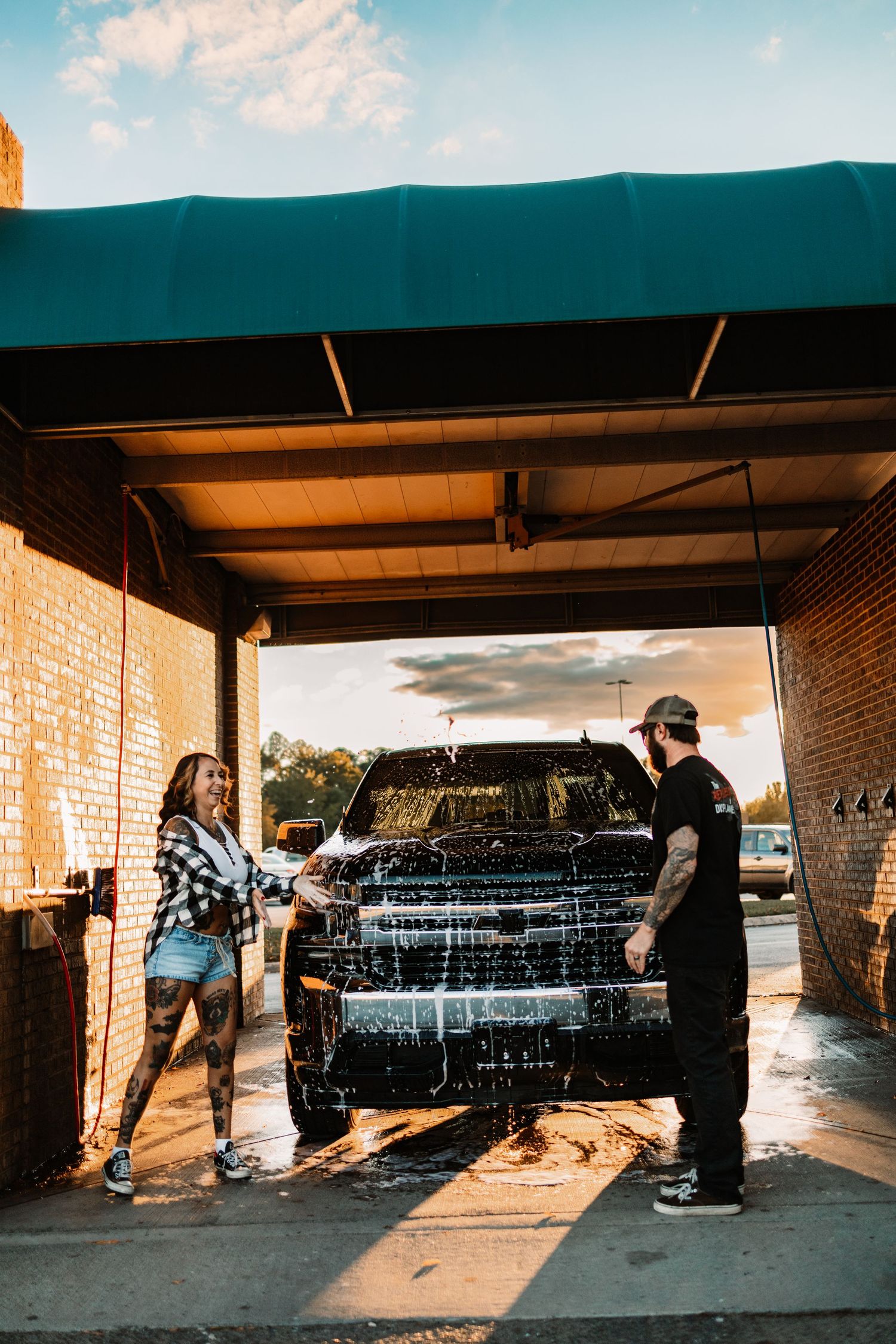 Tattooed couple laughing together while washing a foam-covered black pickup truck at an outdoor car wash bay.