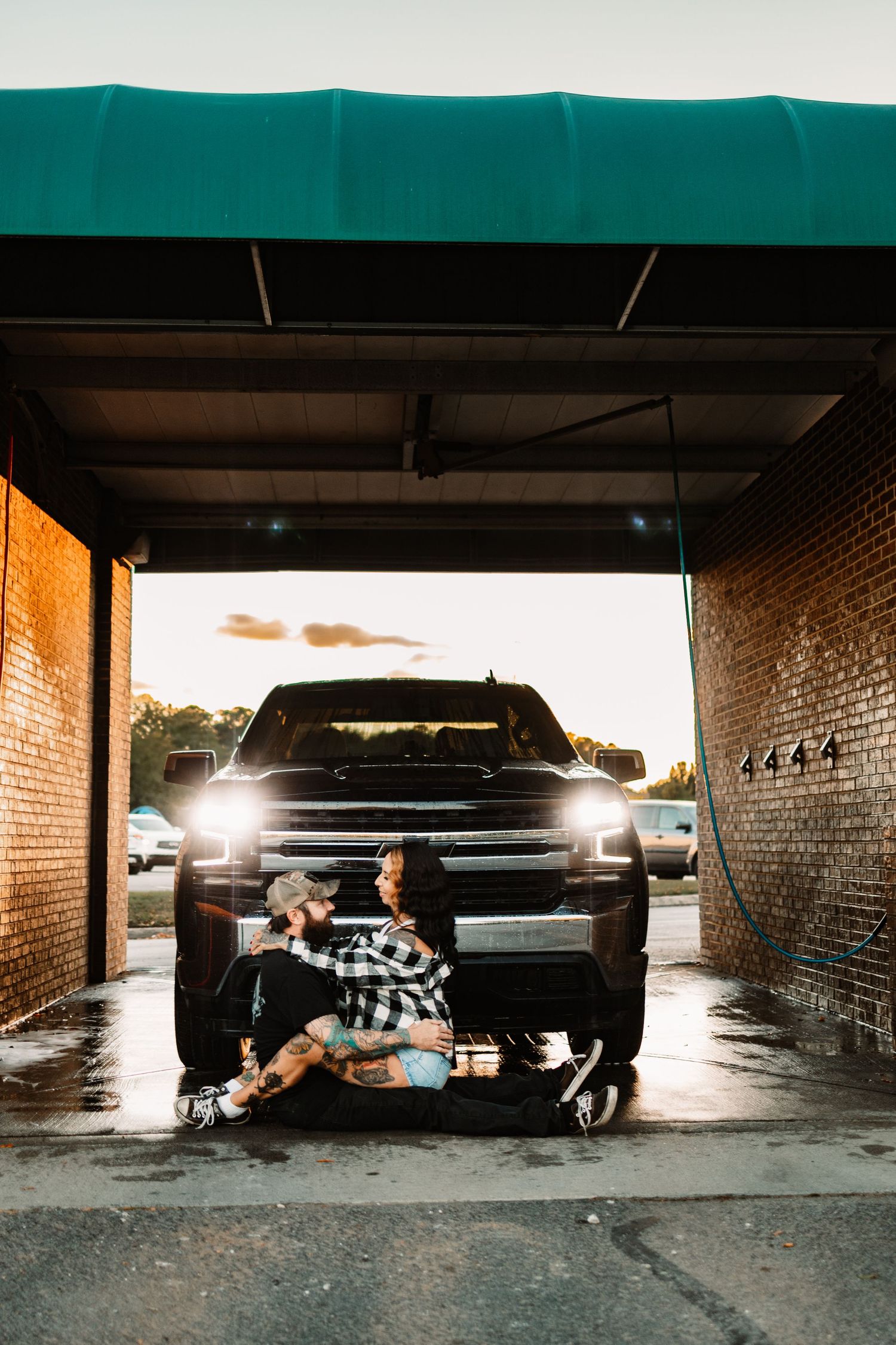 Couple sitting together on wet ground in front of black truck with headlights on at car wash.
