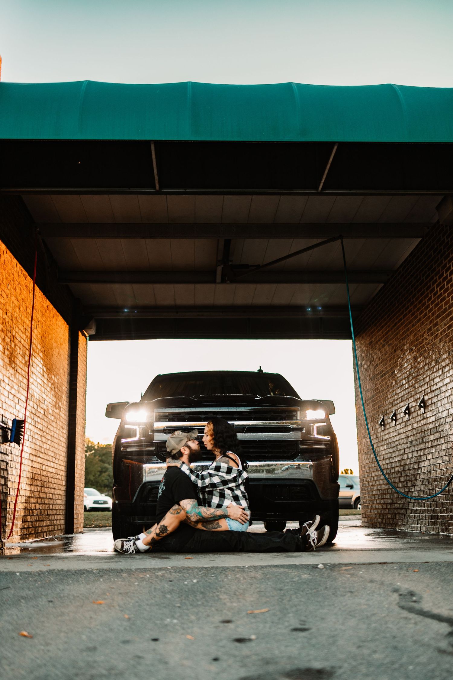 Tattooed couple sitting close together in front of black pickup truck at car wash during golden hour.