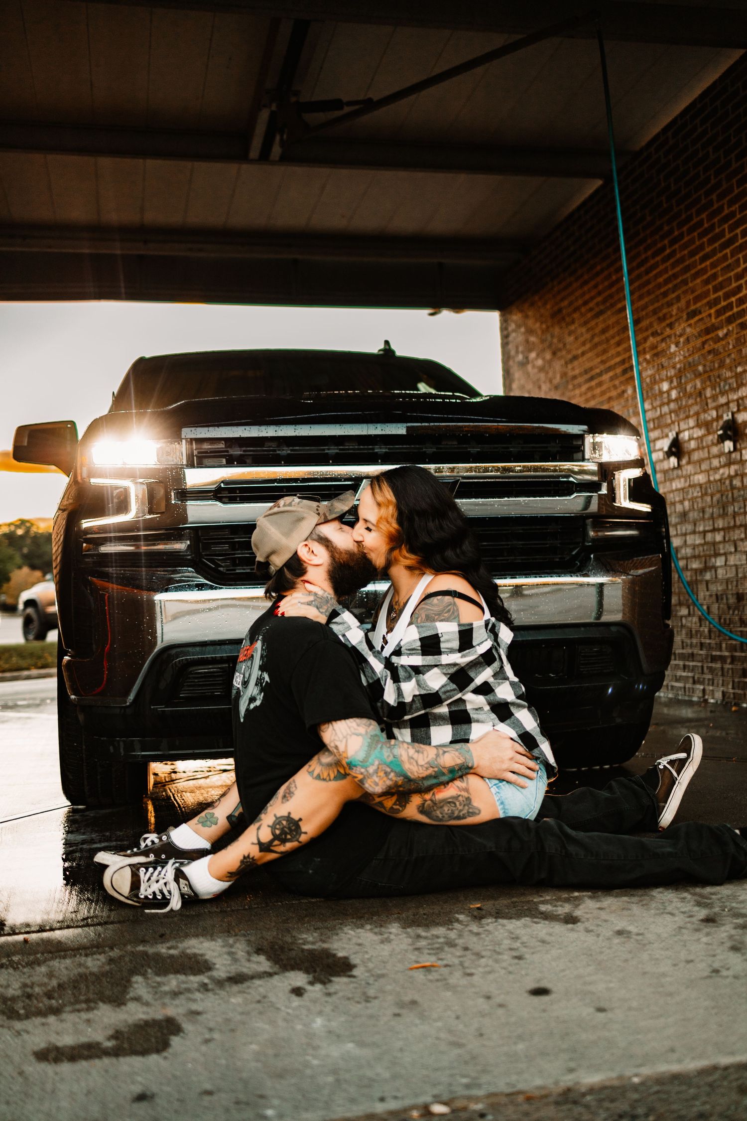 Tattooed couple kissing in front of black Chevrolet truck at car wash during warm sunset light.