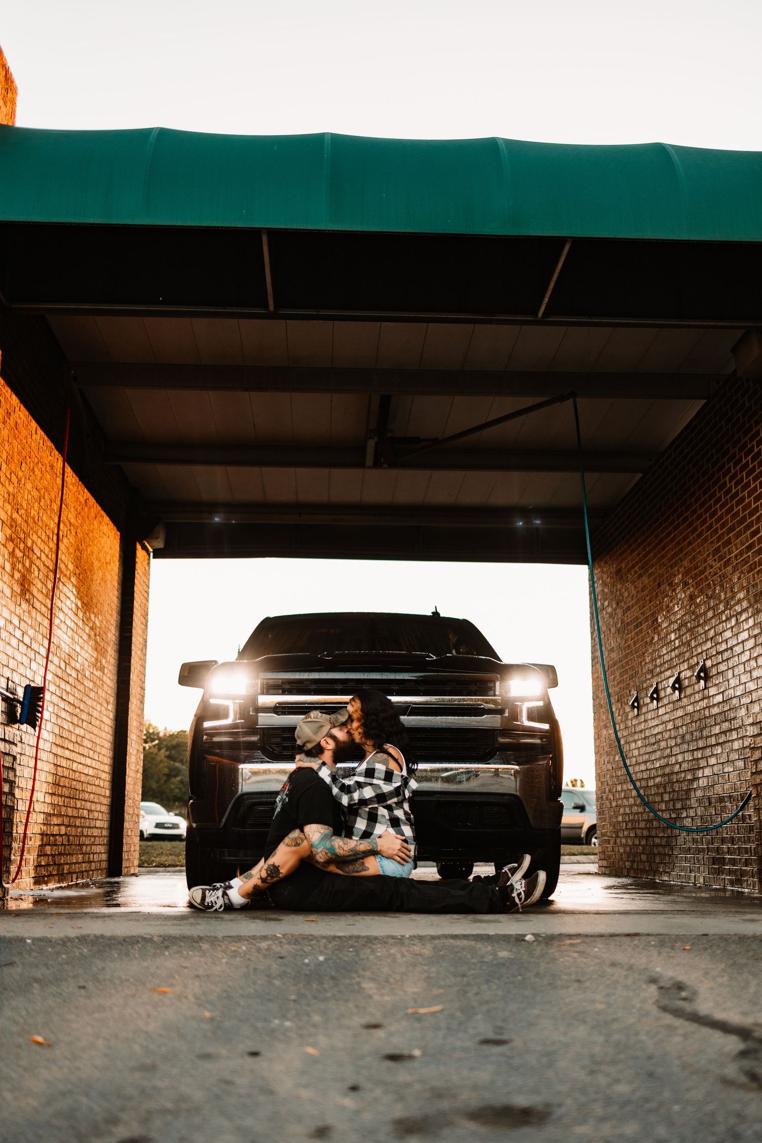 Couple sitting on ground embracing in front of black truck with headlights on at car wash bay.