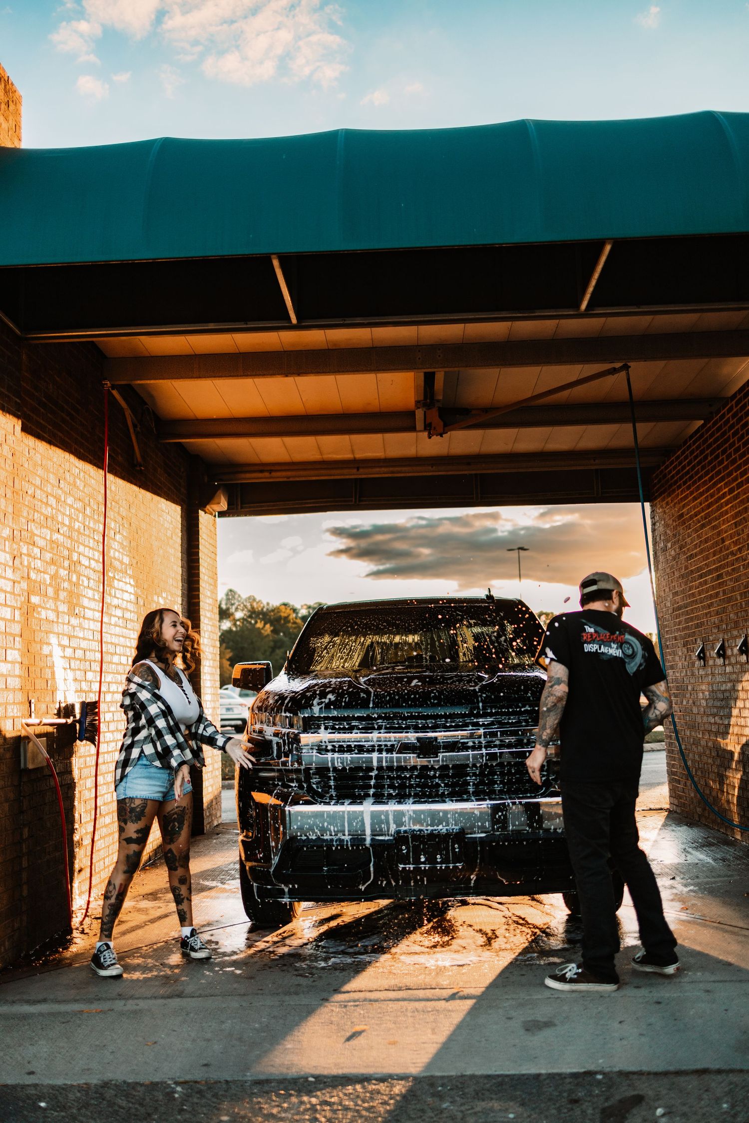 Man and woman with tattoos washing a soapy black truck together at a self-service car wash during golden hour.