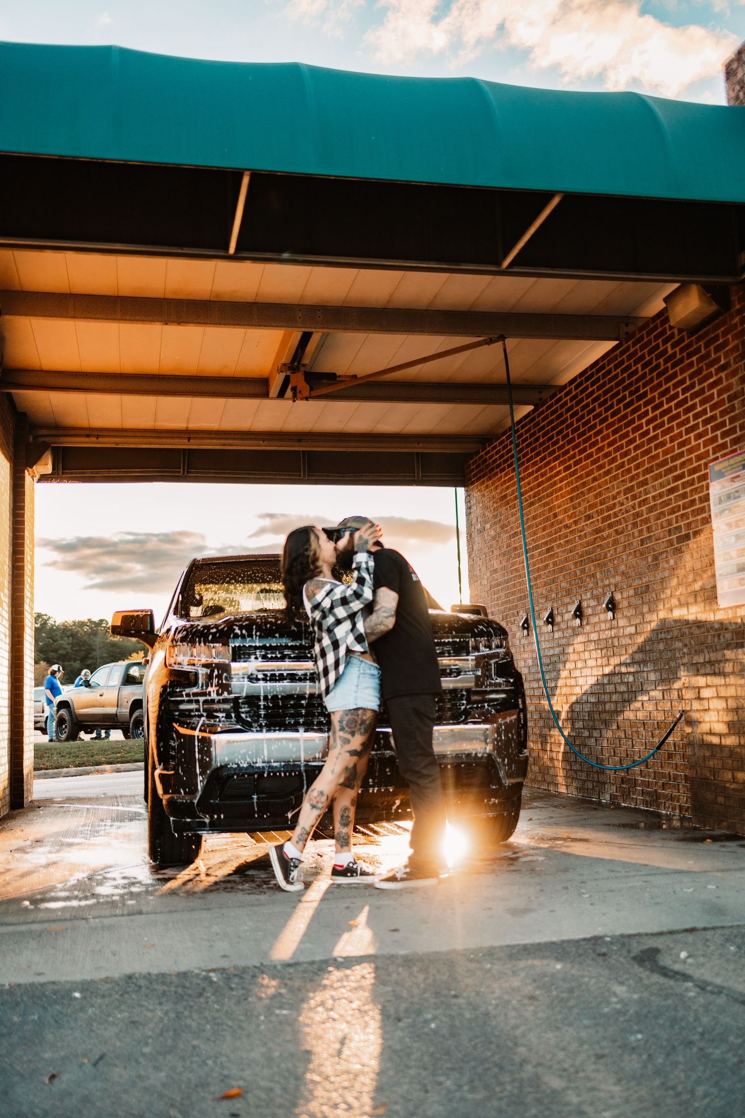 Tattooed couple kissing passionately in front of a sudsy black pickup truck at a brick car wash at sunset.