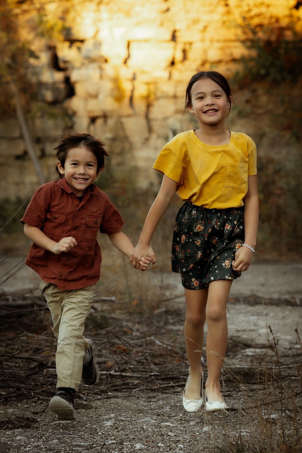 Two children holding hands and playing together outdoors in casual attire against a textured wall background.
