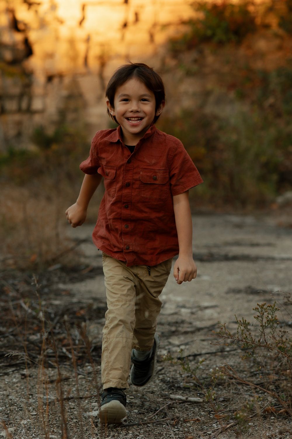 A joyful child wearing a rust-colored shirt and khaki pants runs along a dirt path during golden hour.