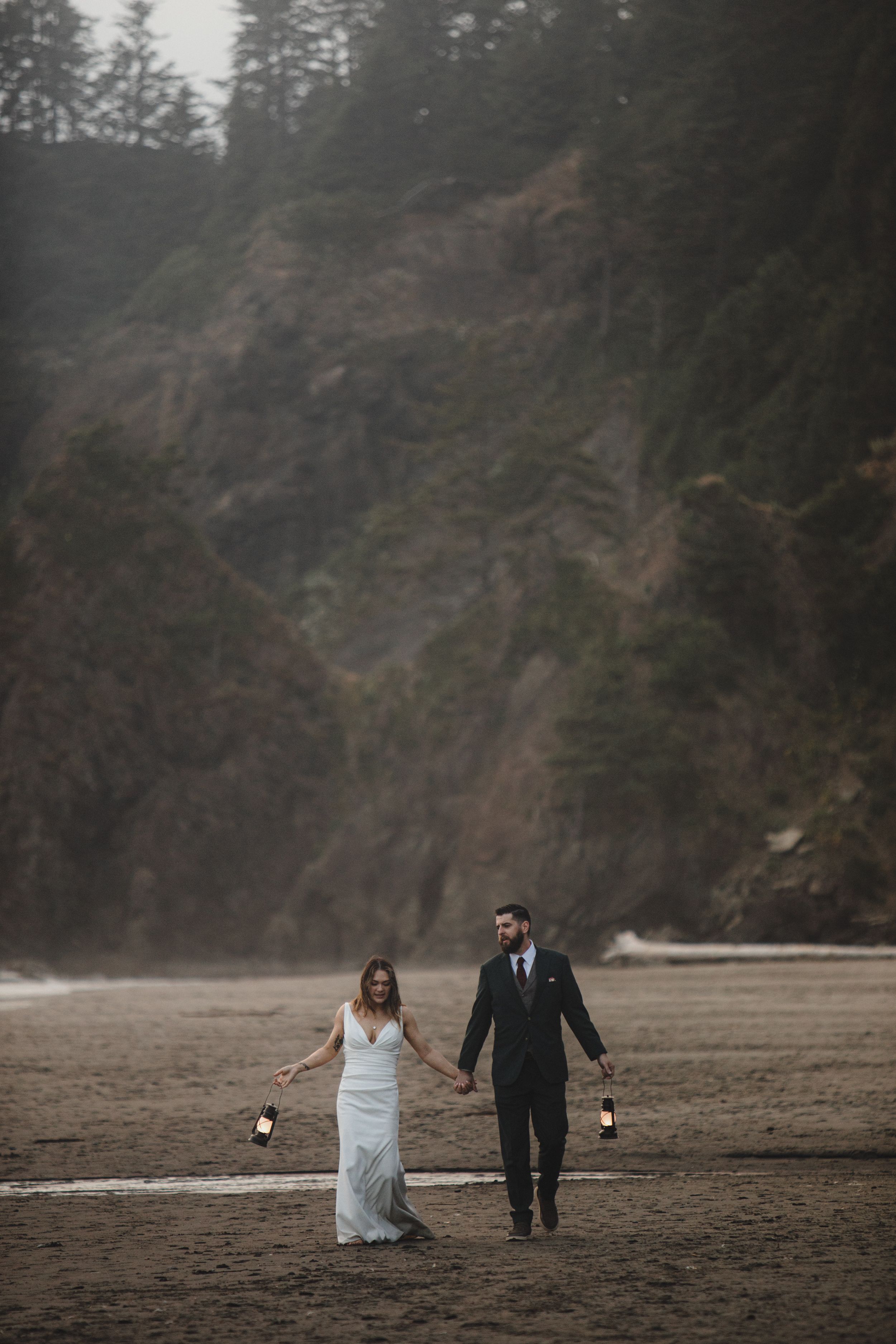 A couple in wedding attire walks hand in hand along a moody Pacific Northwest beach with tall evergreens in background.