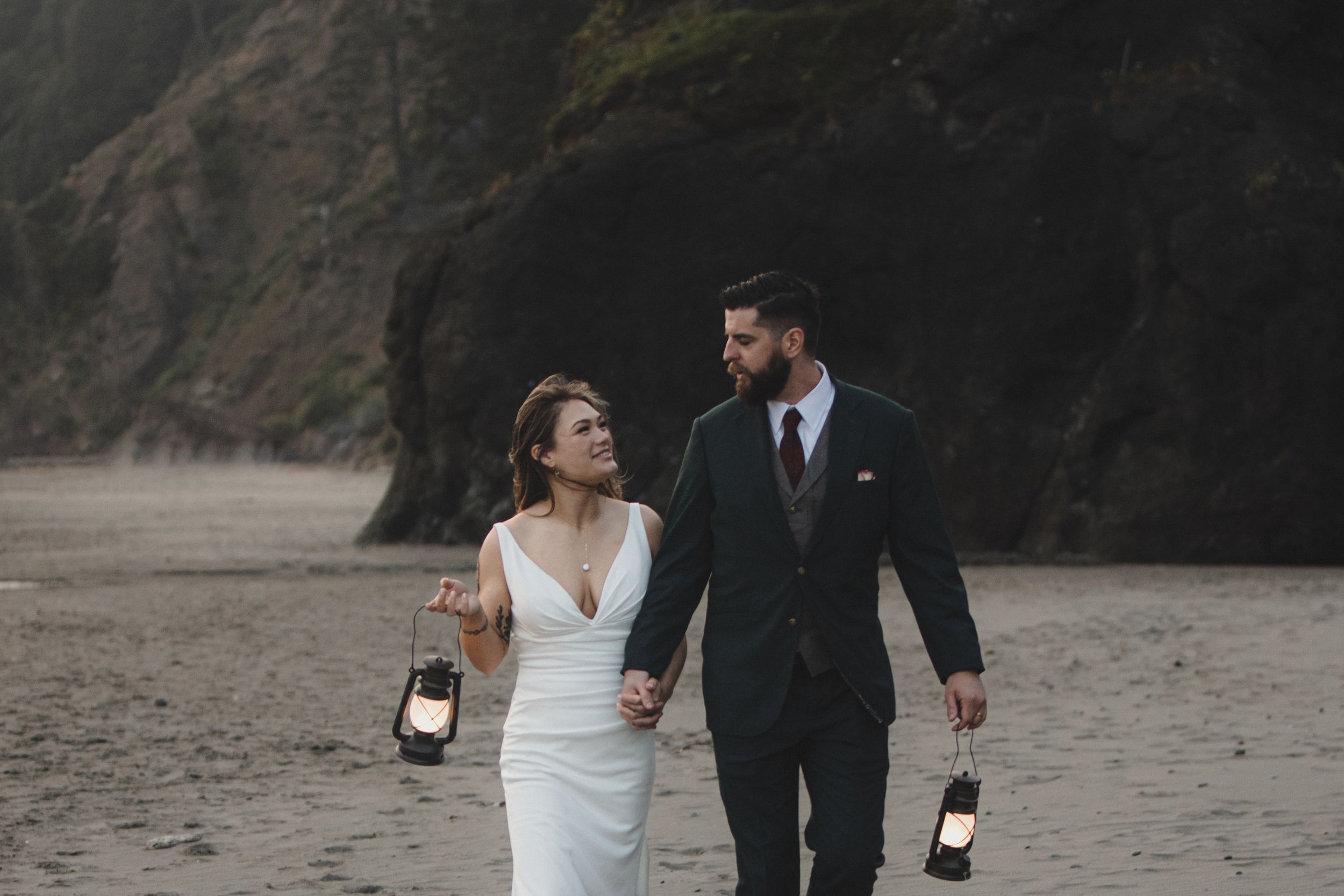 A couple in wedding attire walks along a moody beach at dusk holding vintage lanterns.