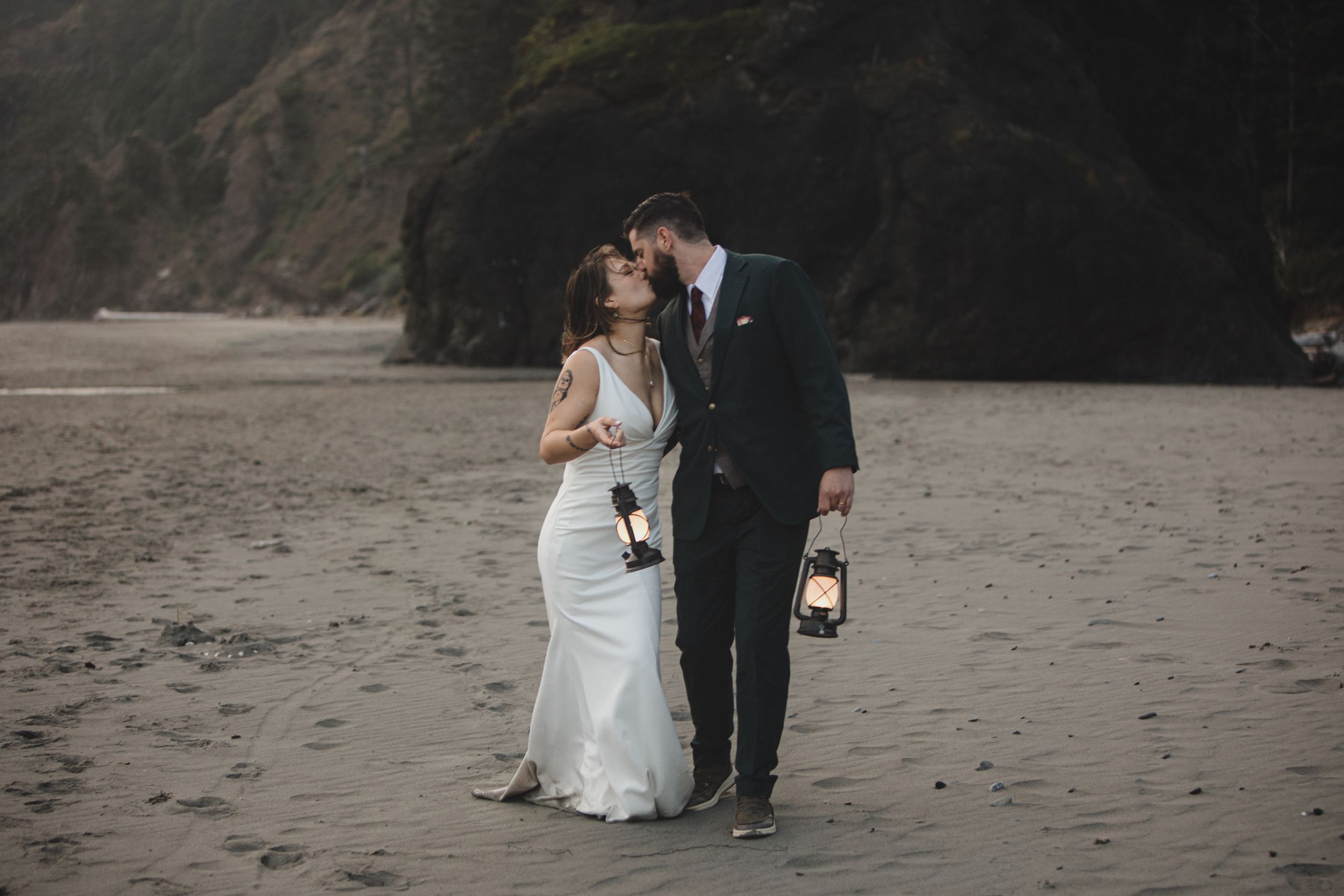 Newlywed couple walks along scenic rocky beach in formal wedding attire at sunset.