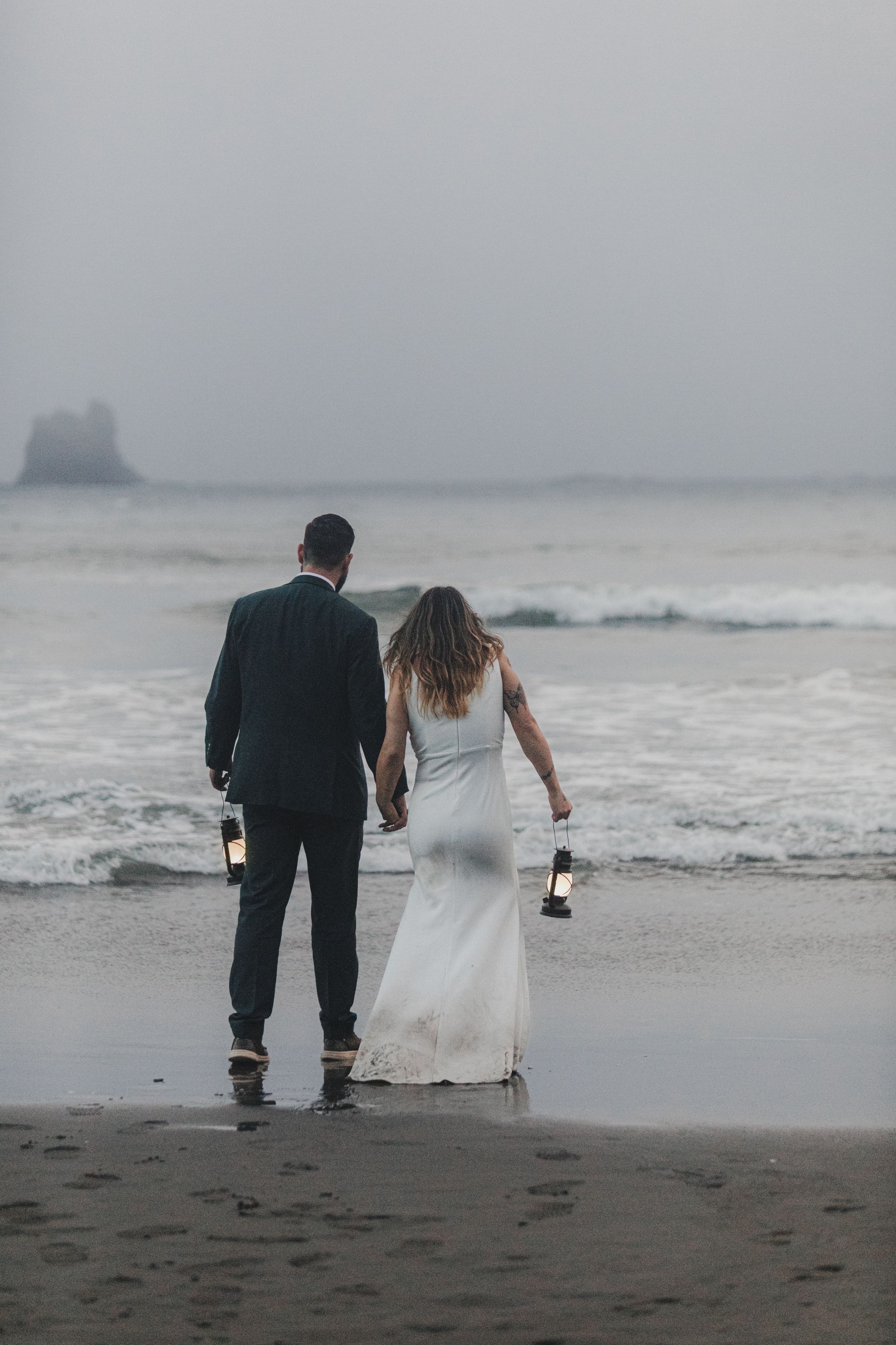 A couple in wedding attire holds hands while standing on a misty beach at dusk with waves crashing in the background.