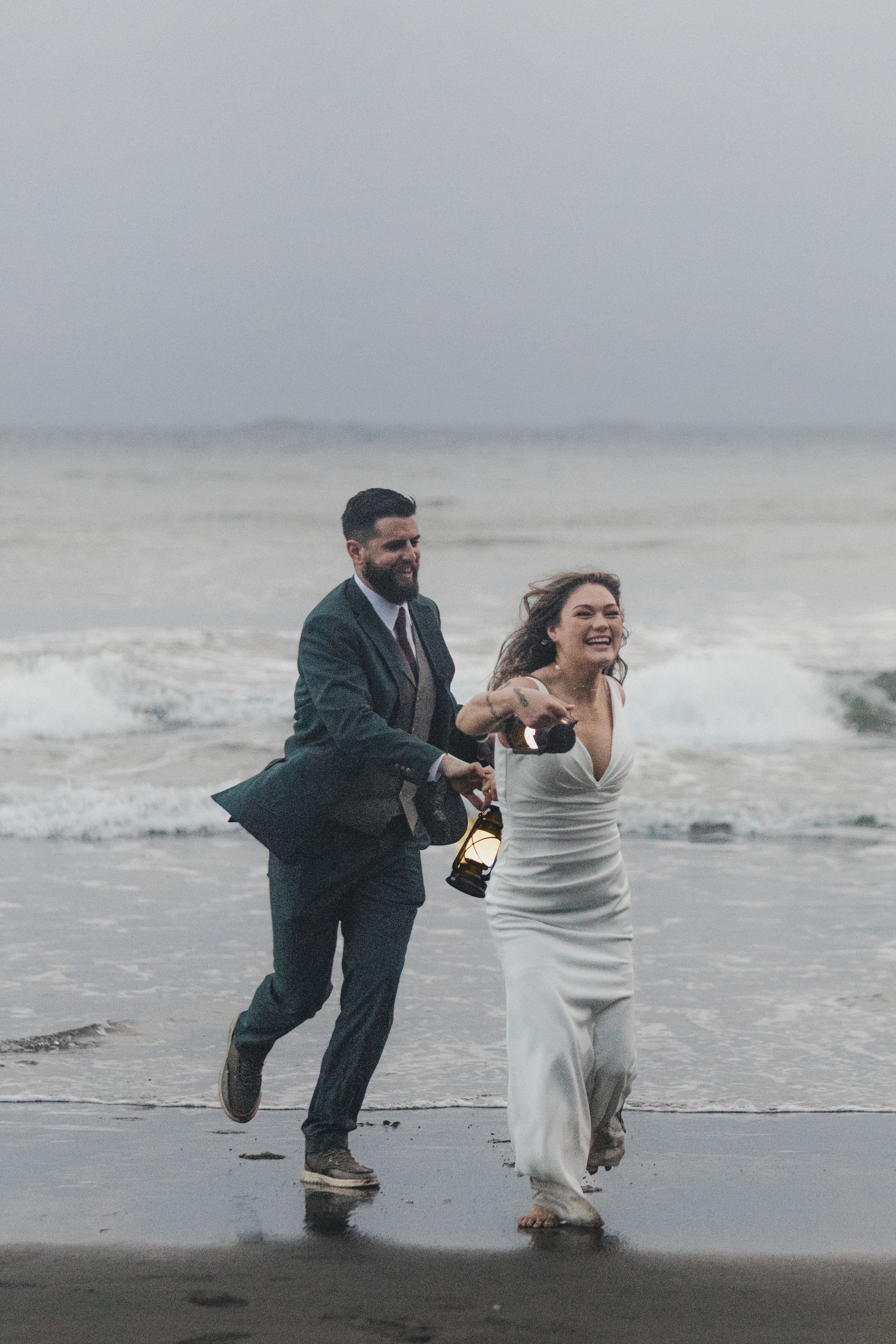 A couple in wedding attire runs joyfully along a misty beach while holding a champagne bottle.