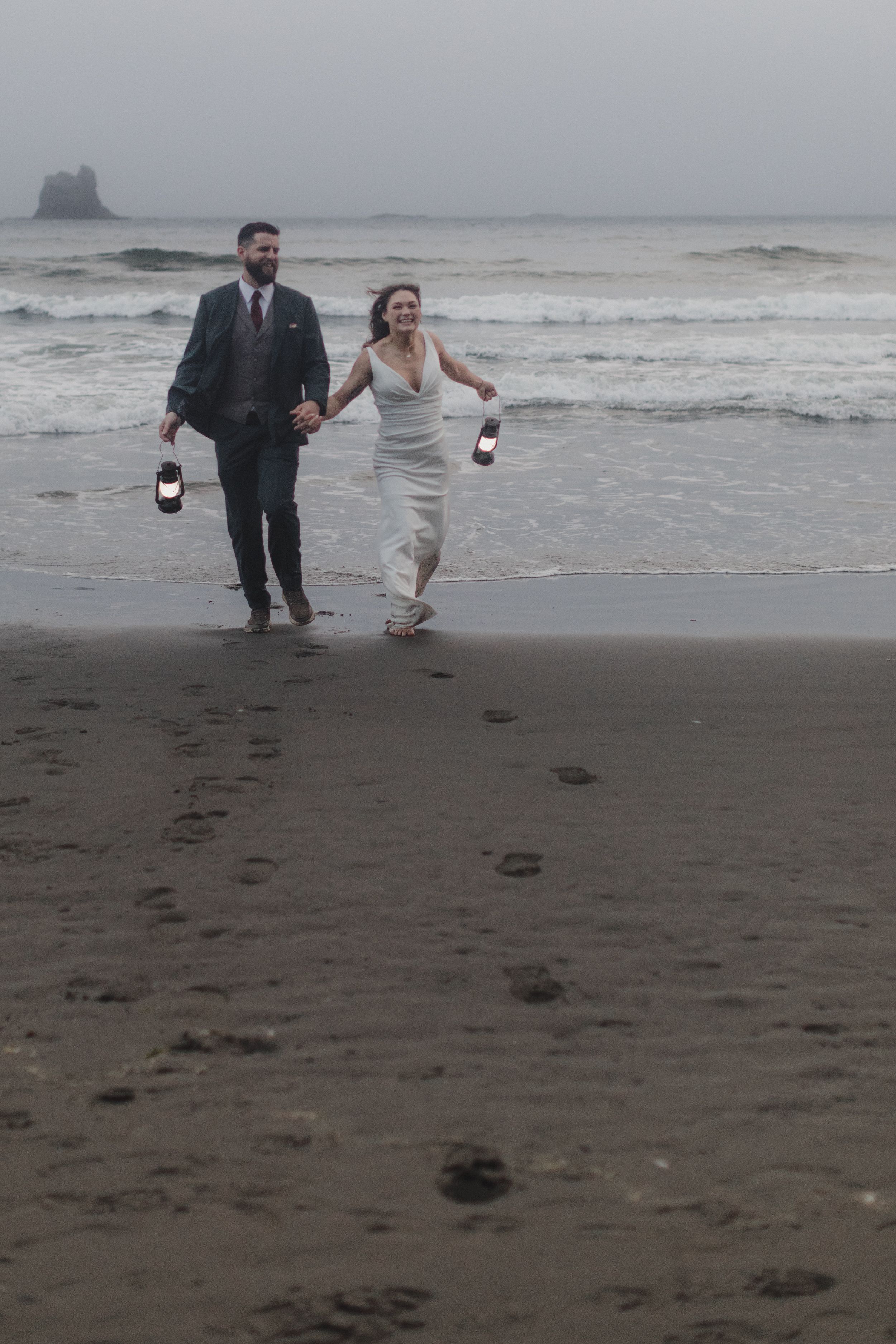 A couple in wedding attire runs joyfully along a misty beach holding wine bottles on their special day.