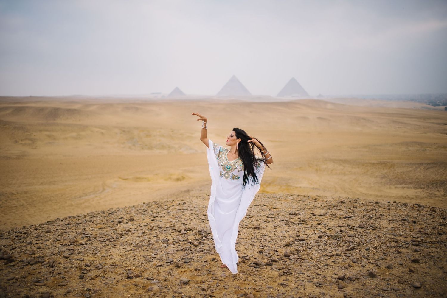 A flowing white dress catches the wind against the backdrop of Egyptian pyramids in the desert.