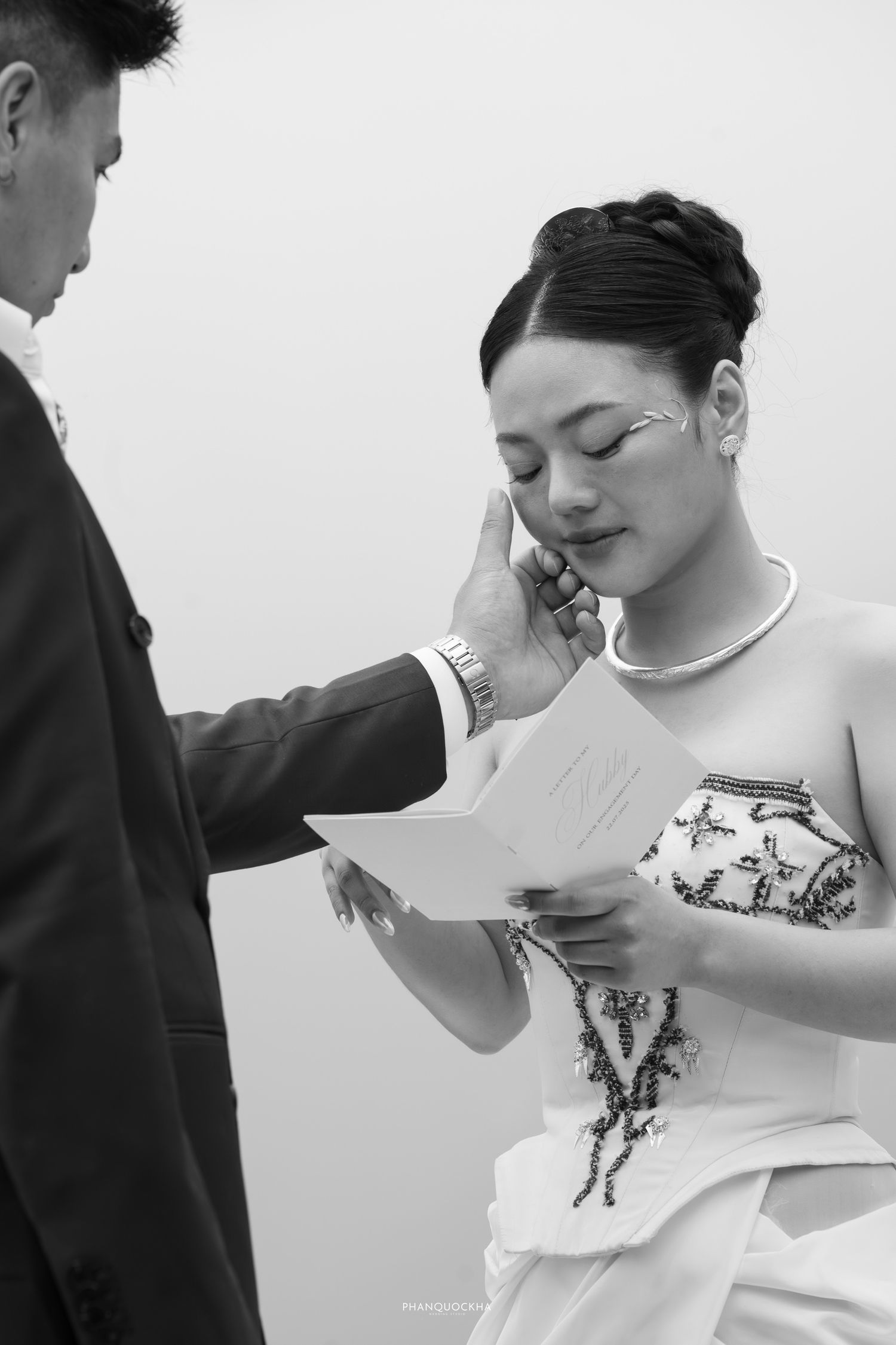 Emotional bride in embroidered dress reads vows as groom gently wipes her tears in black and white.