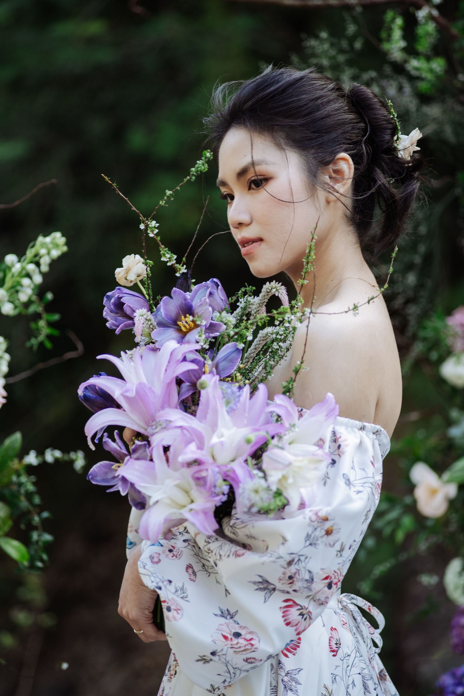 Young Asian woman in floral off-shoulder gown holds purple lily bouquet outdoors.