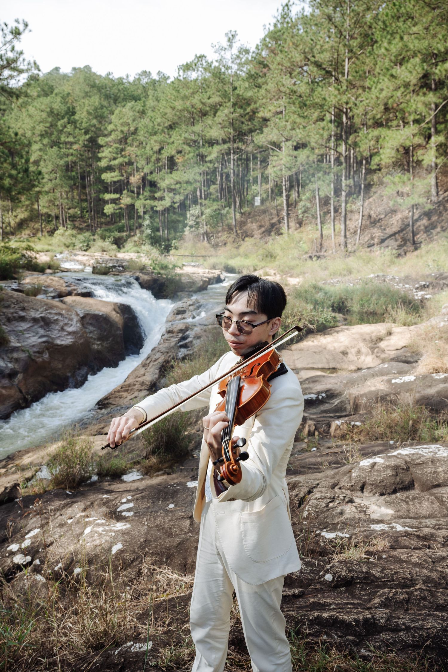 Man in white suit plays violin near rocky waterfall in pine forest.