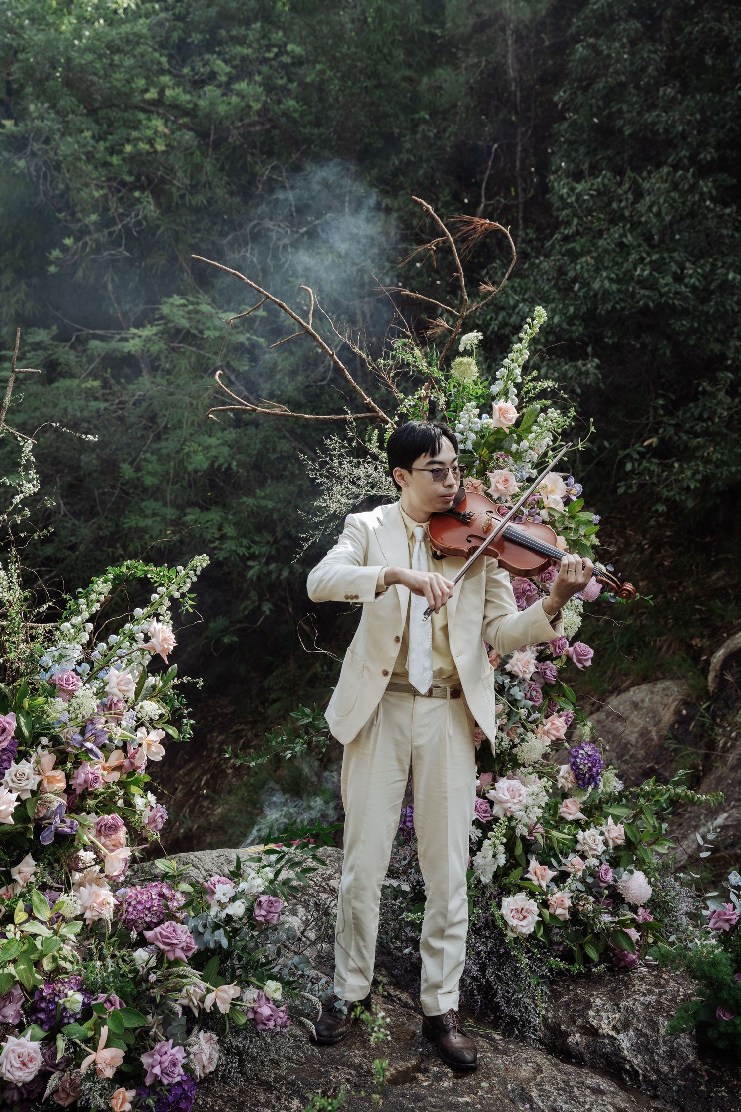 Violinist in cream suit performs amid lush floral arrangements in misty forest.