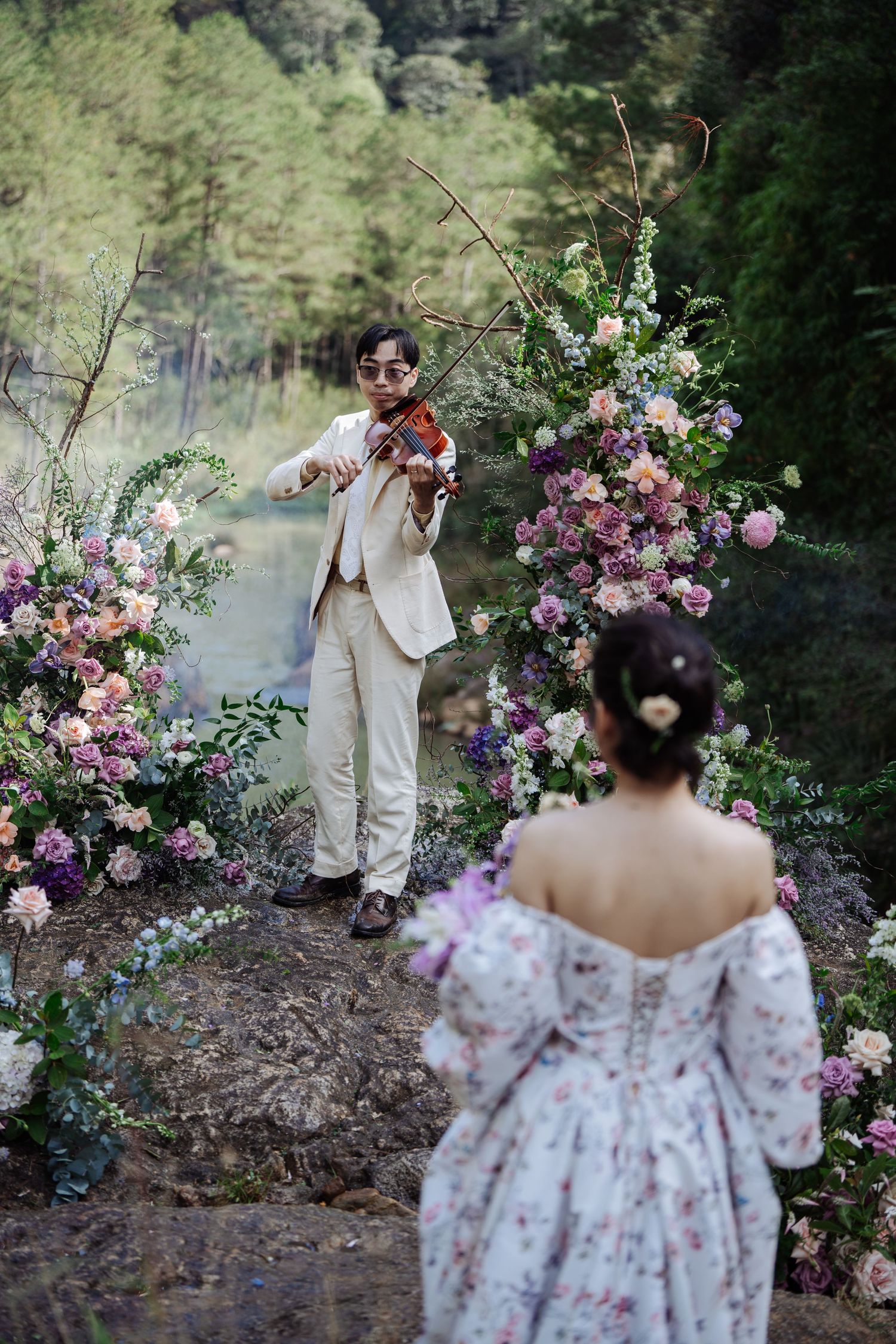 Groom plays violin for bride among colorful flowers by forest lake.