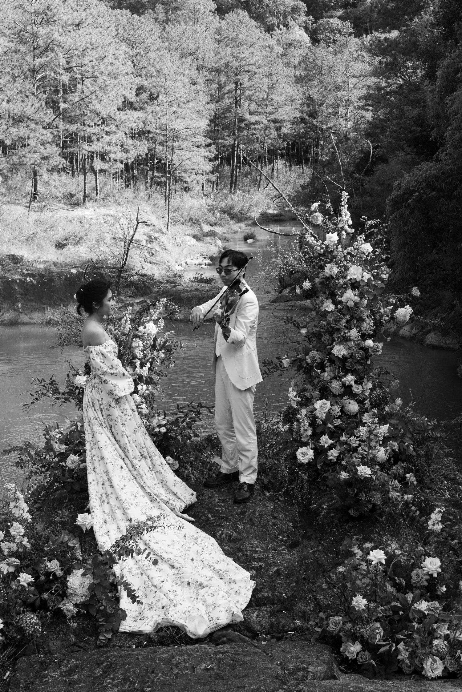 Black and white photo of couple exchanging vows by river with floral arch.