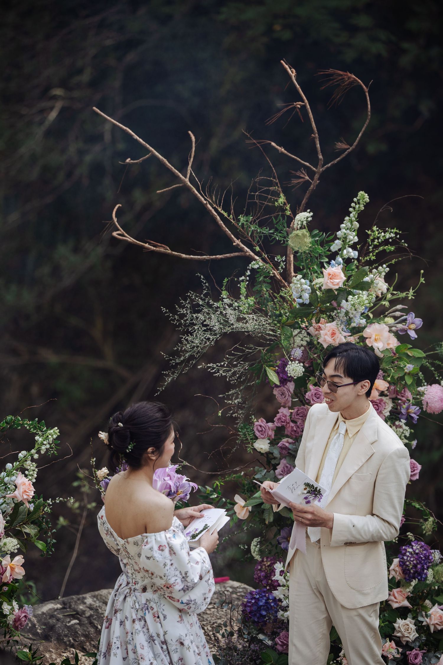 Couple reads wedding vows under floral arch with twisted branches in forest.