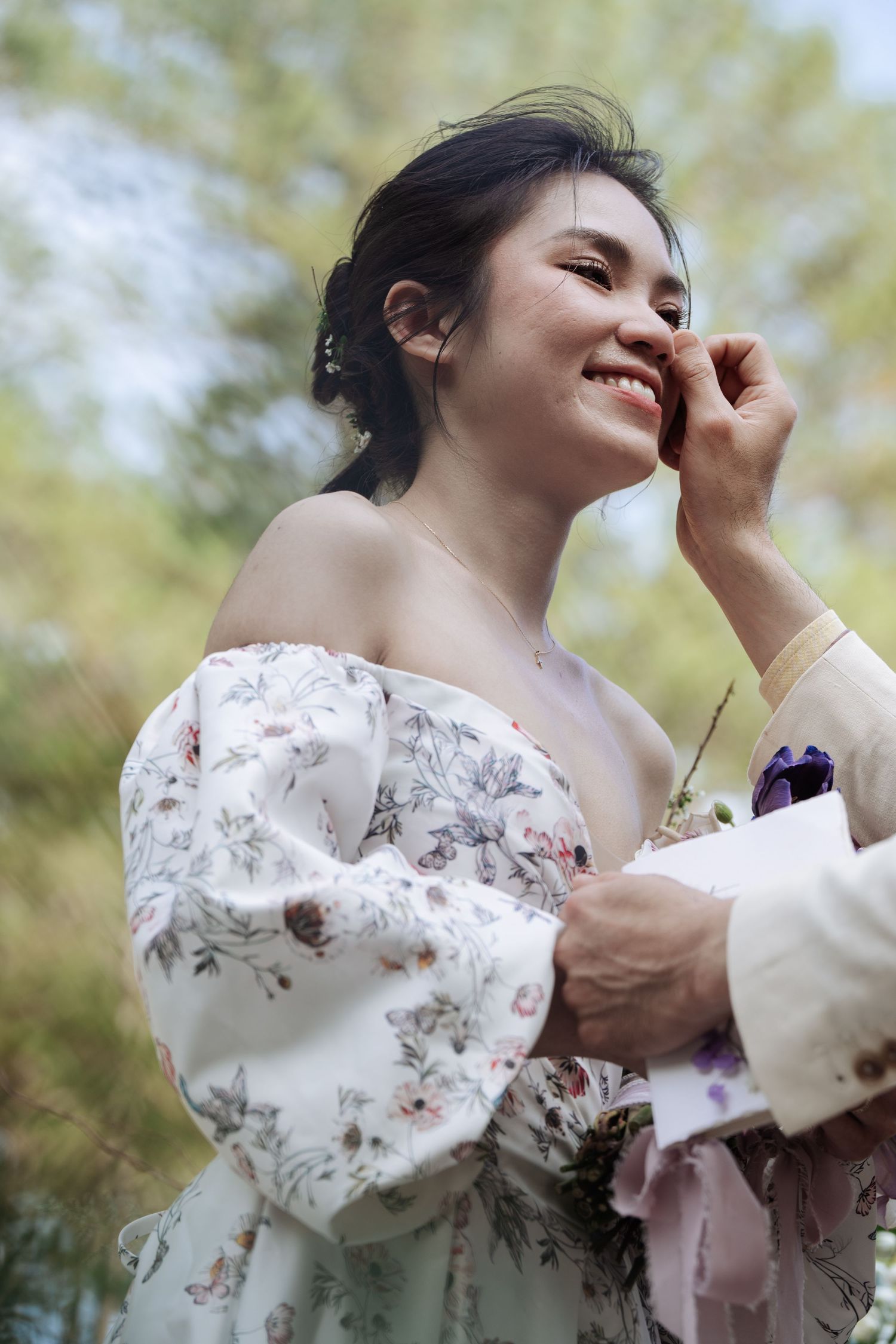 Smiling bride in floral off-shoulder gown touches her face during outdoor wedding ceremony.