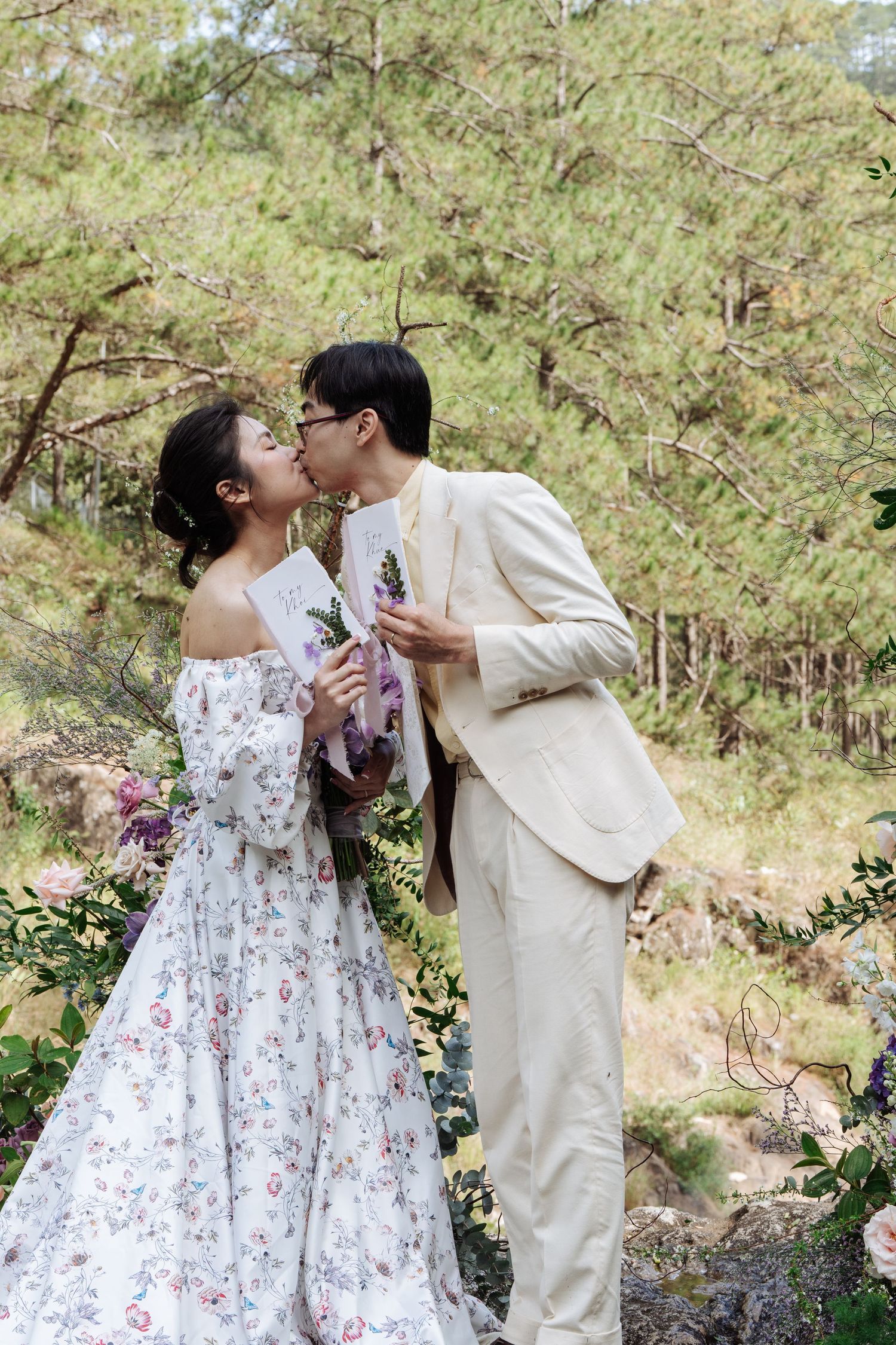 Couple sharing a kiss in nature, bride in floral gown and groom in cream suit holding flowers.