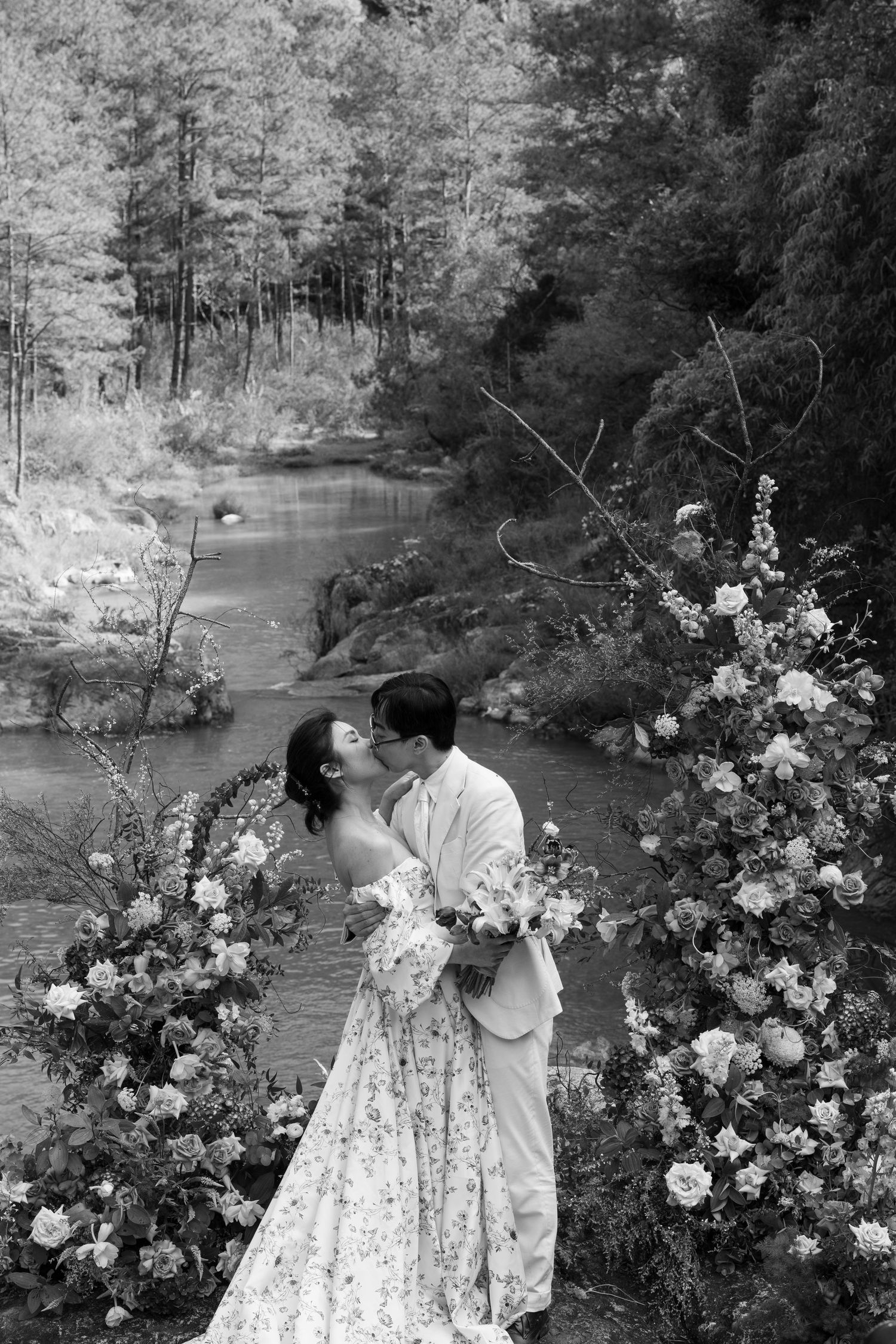Black and white photo of couple kissing by a stream framed by elegant floral arrangements.