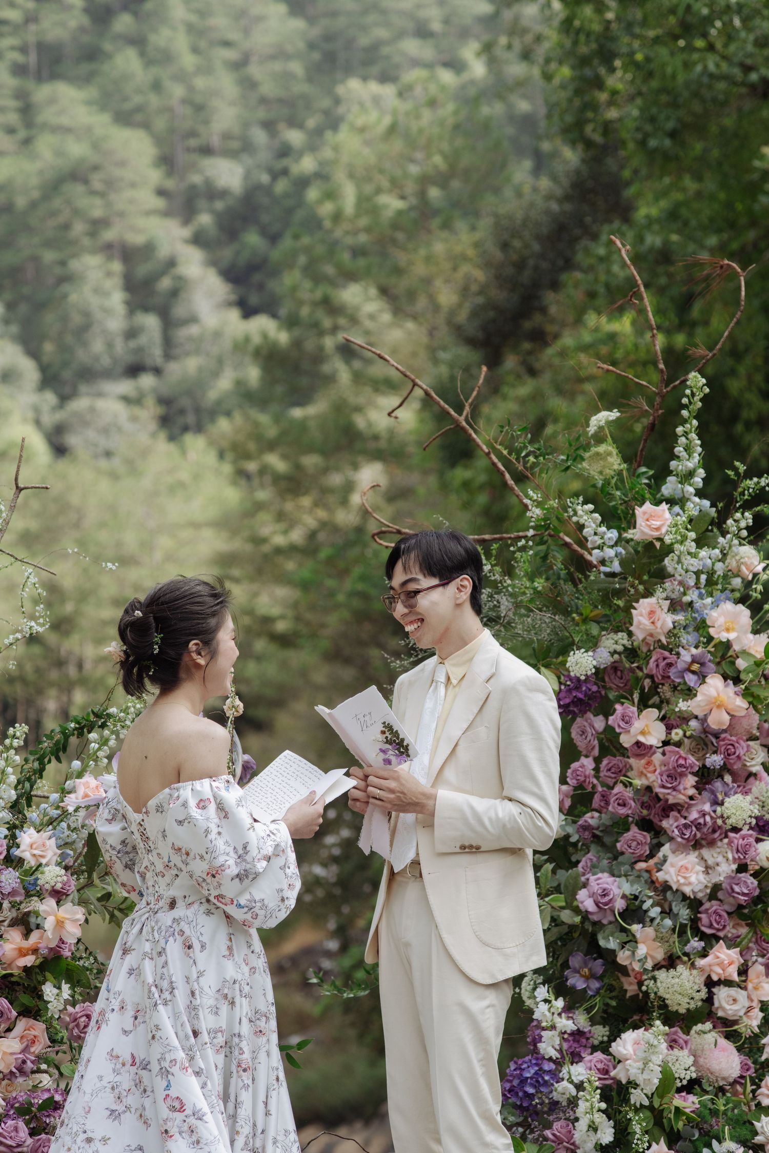 Bride and groom exchange vows surrounded by lush floral arrangements in a forest setting.