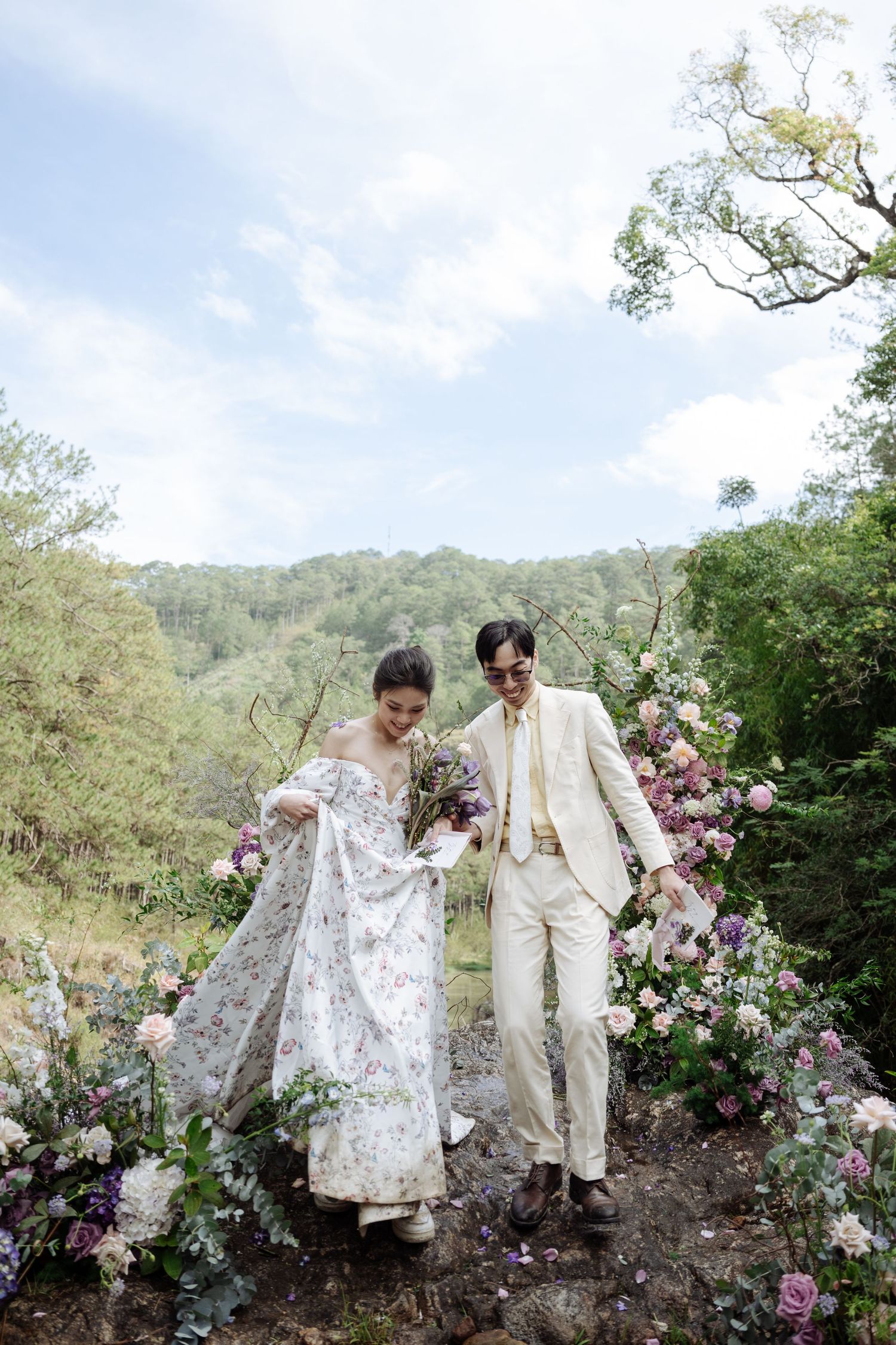 Bride and groom walk together through floral arch in scenic outdoor forest wedding setting.