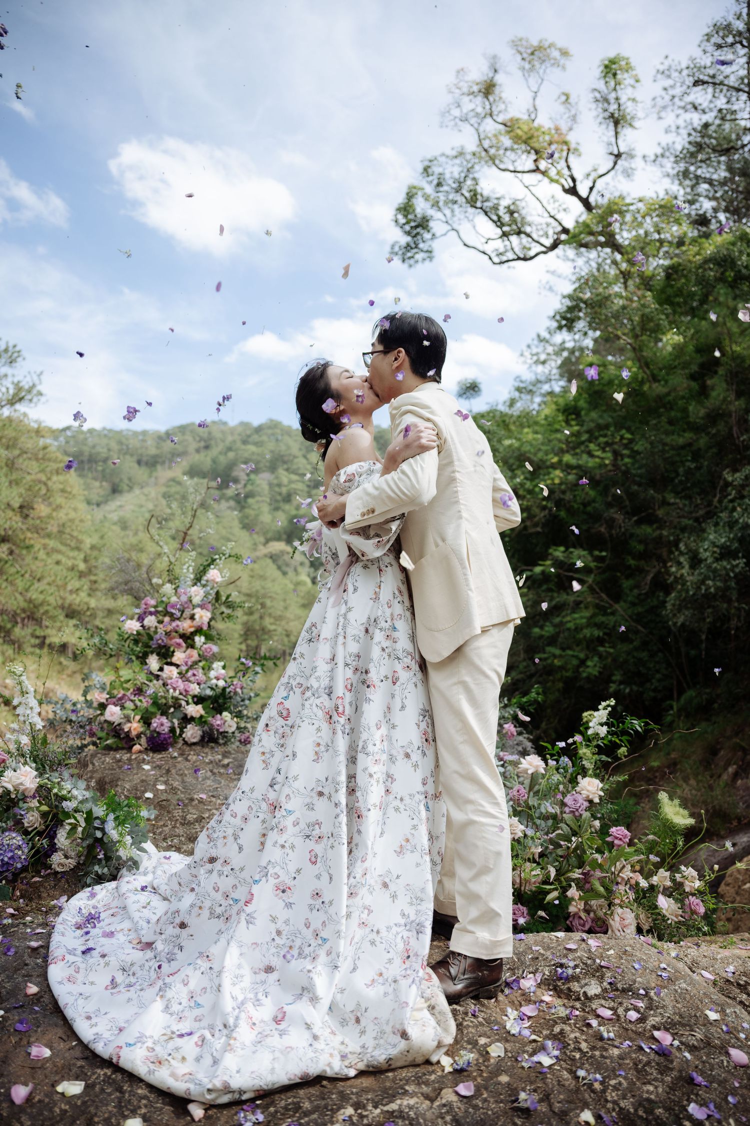 Couple kisses as flower petals fall around them at outdoor wedding with floral backdrop.