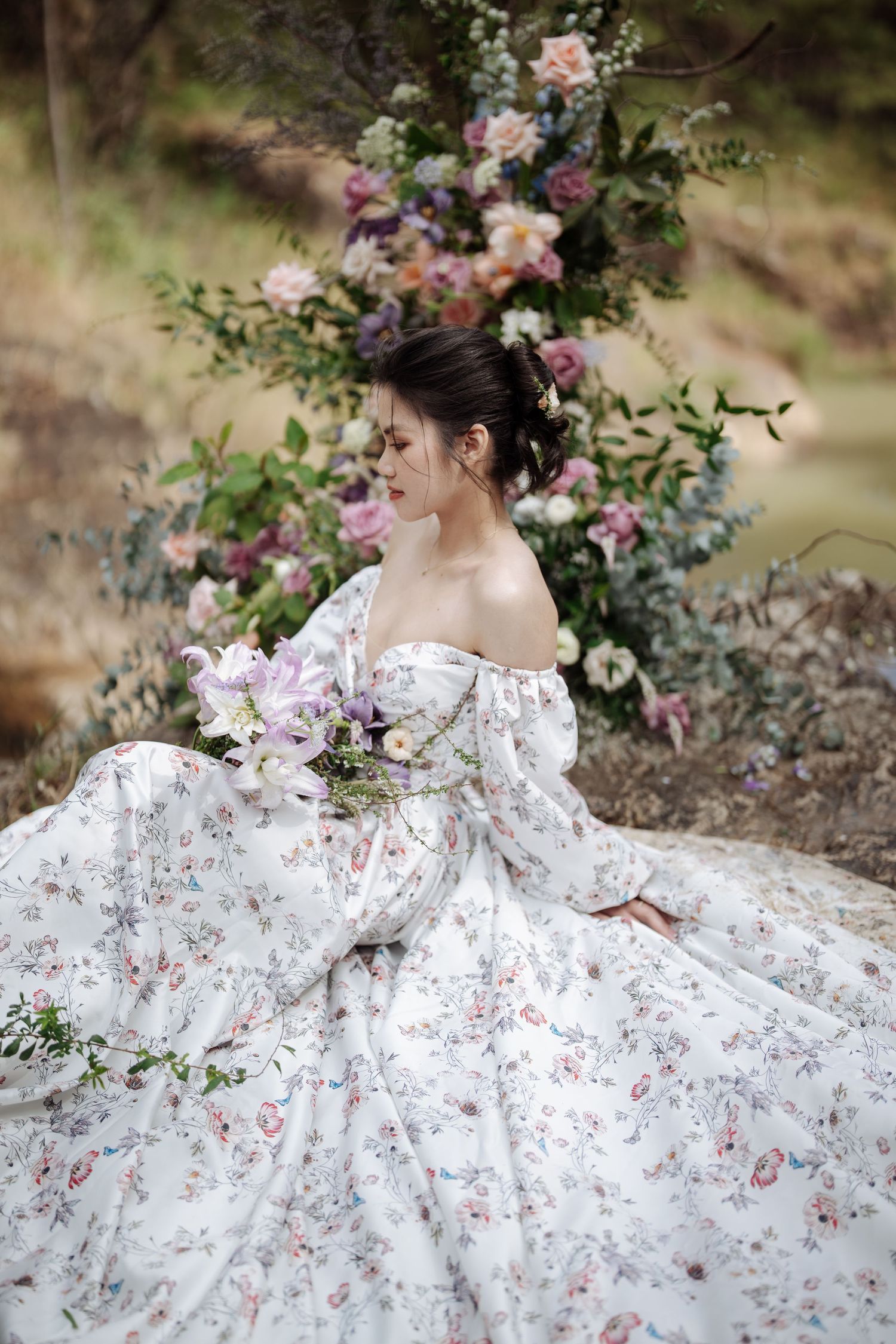 Elegant bride in floral off-shoulder gown sits gracefully before lush pastel floral arrangement.
