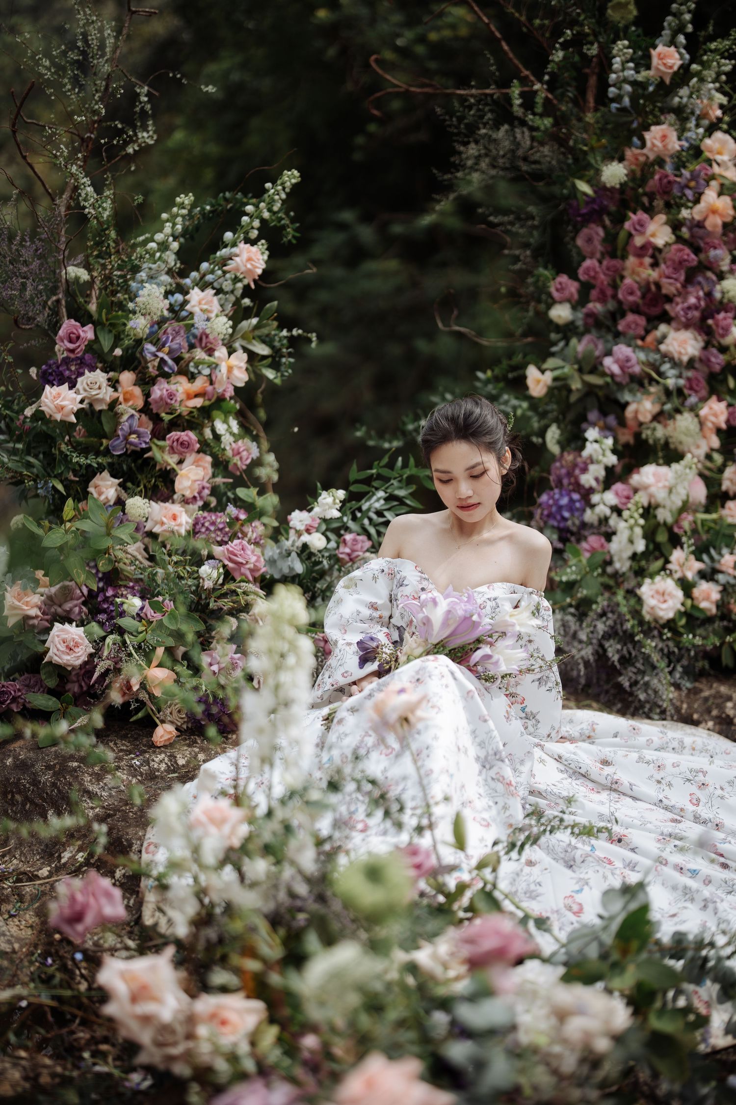 A bride in a floral off-shoulder gown sits surrounded by lush pink and purple flower arrangements in a garden setting.