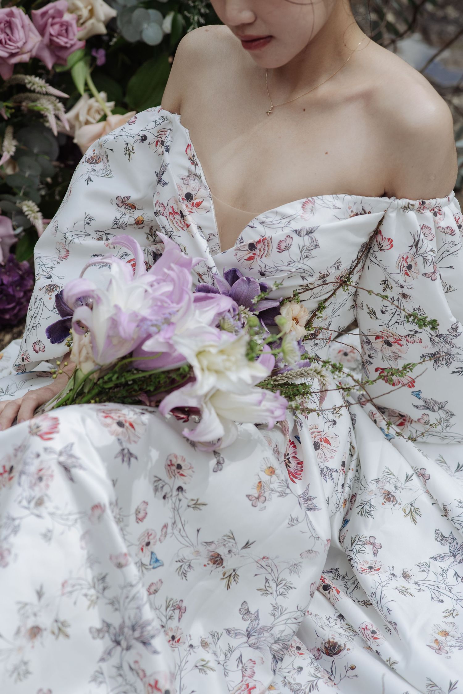 Close-up of a bride in a white floral off-shoulder dress holding a bouquet of purple and white lilies.