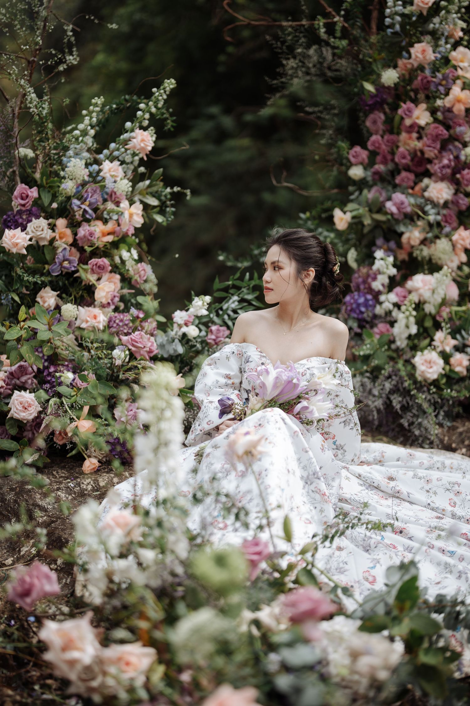 A bride in a white floral ball gown sits gracefully among abundant pink and purple floral arrangements outdoors.