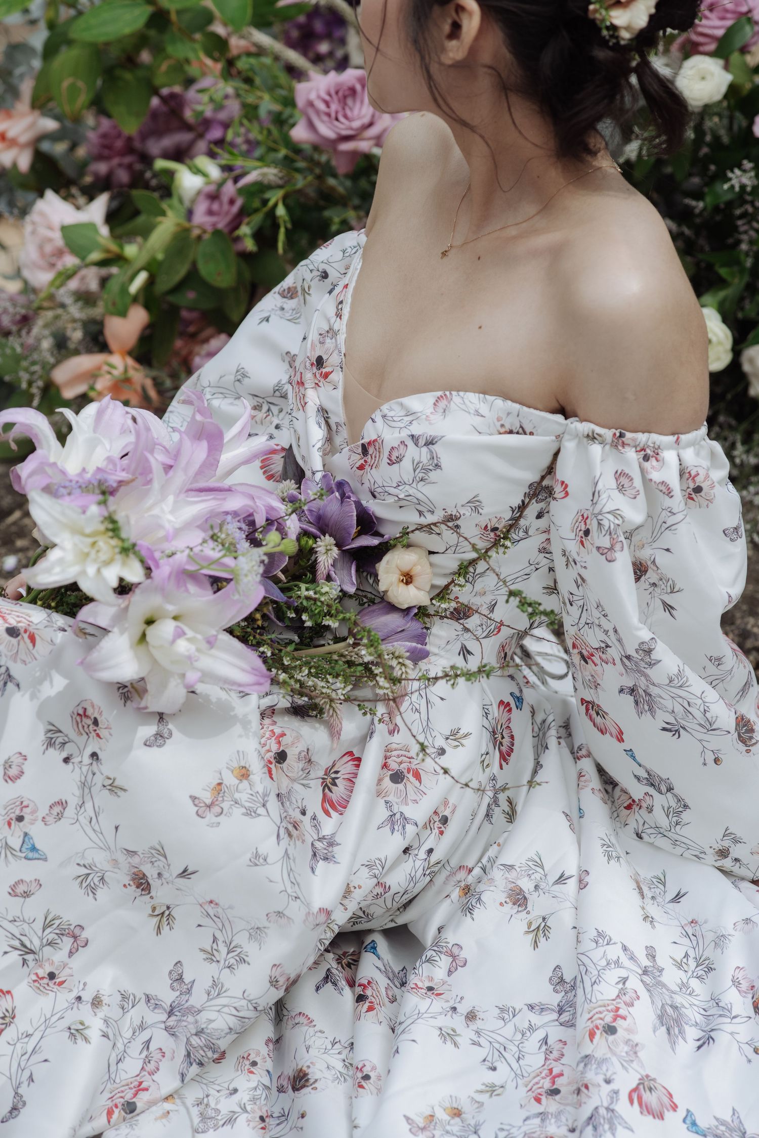 Back view of a bride in a floral off-shoulder gown holding a purple and white lily bouquet outdoors.