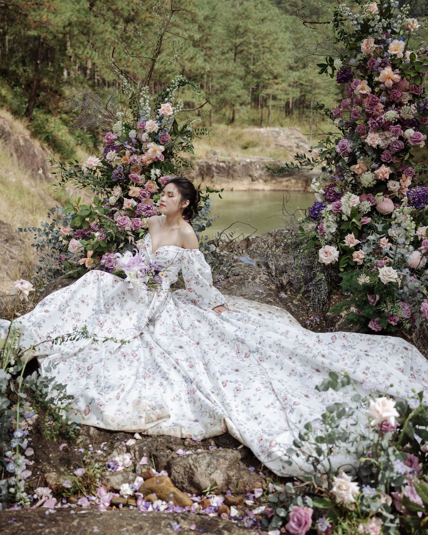 A bride in a flowing white floral gown poses among colorful flower arrangements on rocky terrain near a forest lake.