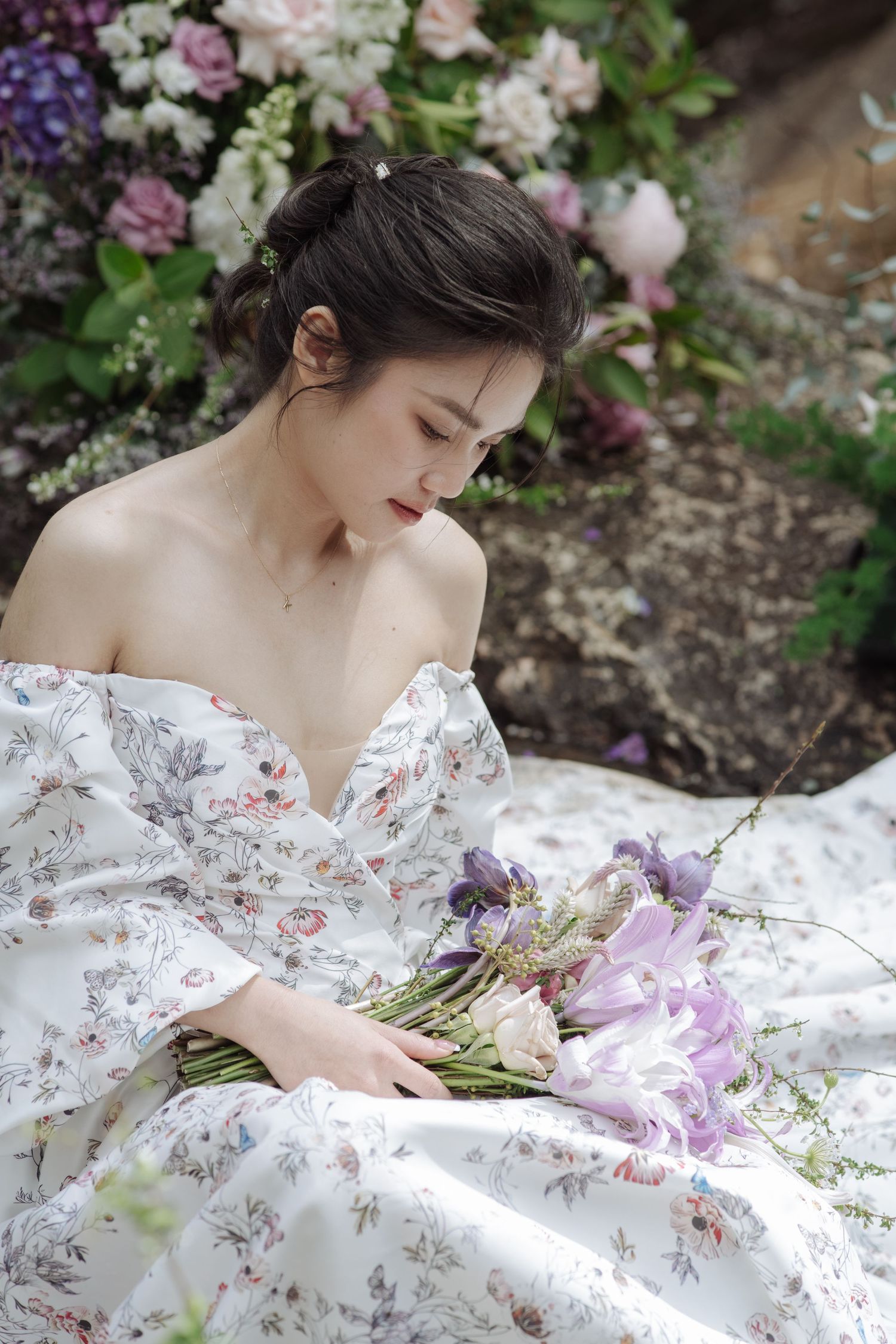 A bride with an updo hairstyle in a floral off-shoulder dress gazes down at her purple lily bouquet.