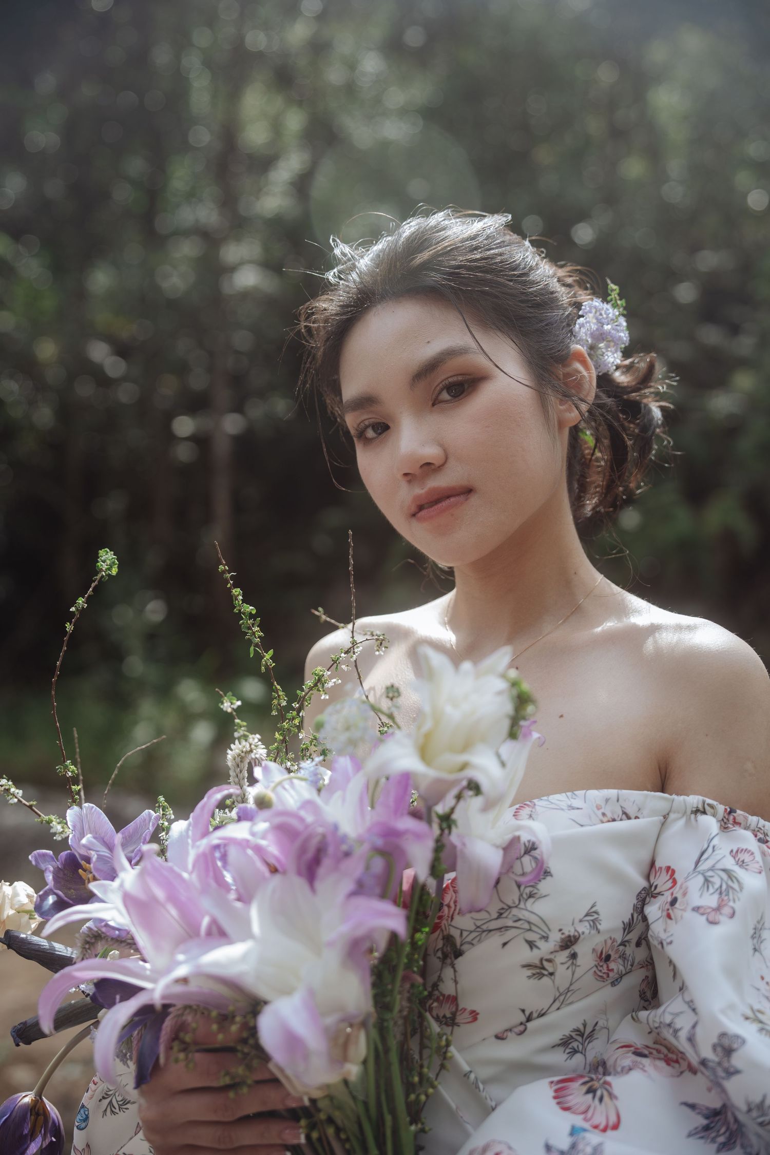A bride in a white floral off-shoulder gown holds a purple and white lily bouquet in a sunlit forest.