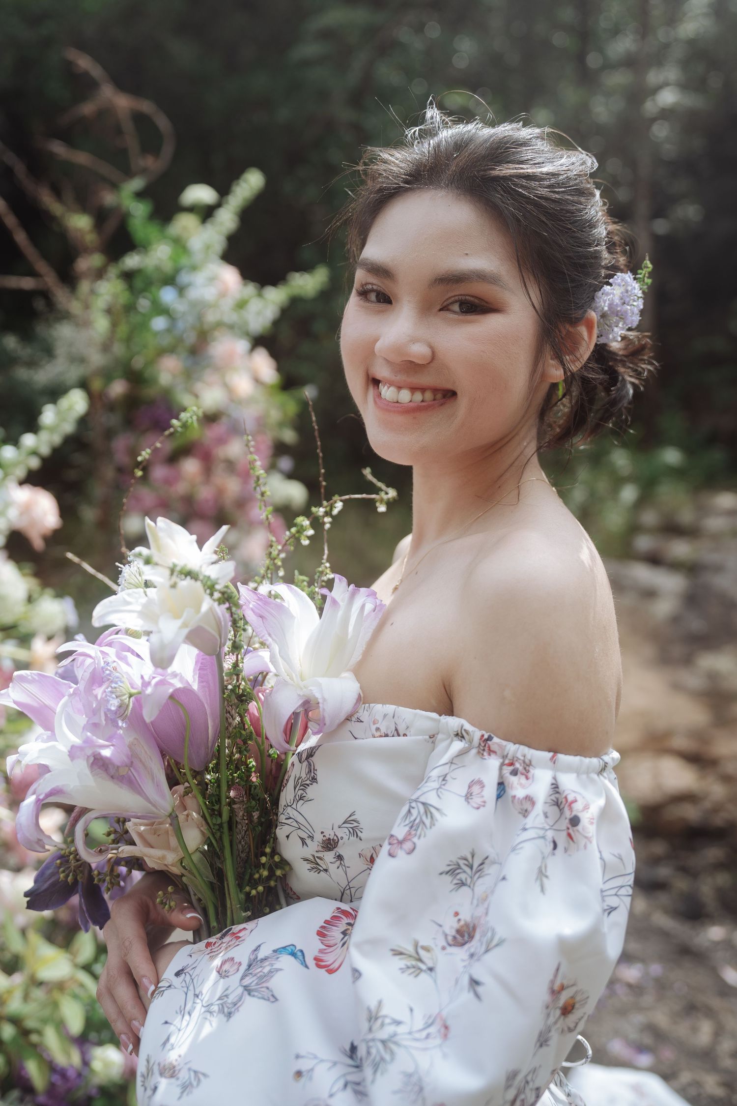 A smiling bride in a white floral off-shoulder dress holds a purple and white lily bouquet in a garden.